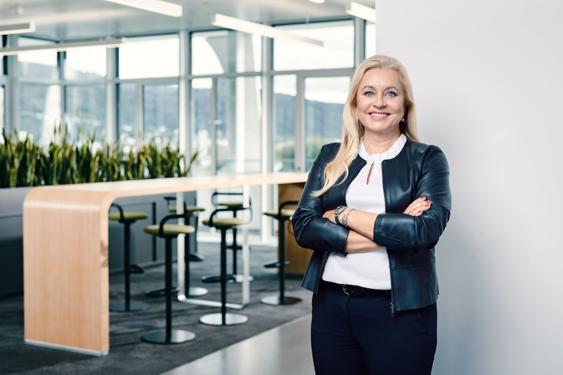 Katharina Beumelburg mit schwarzer Jacke, schwarzer Hose und weißem Oberteil vor einer Fensterfront.