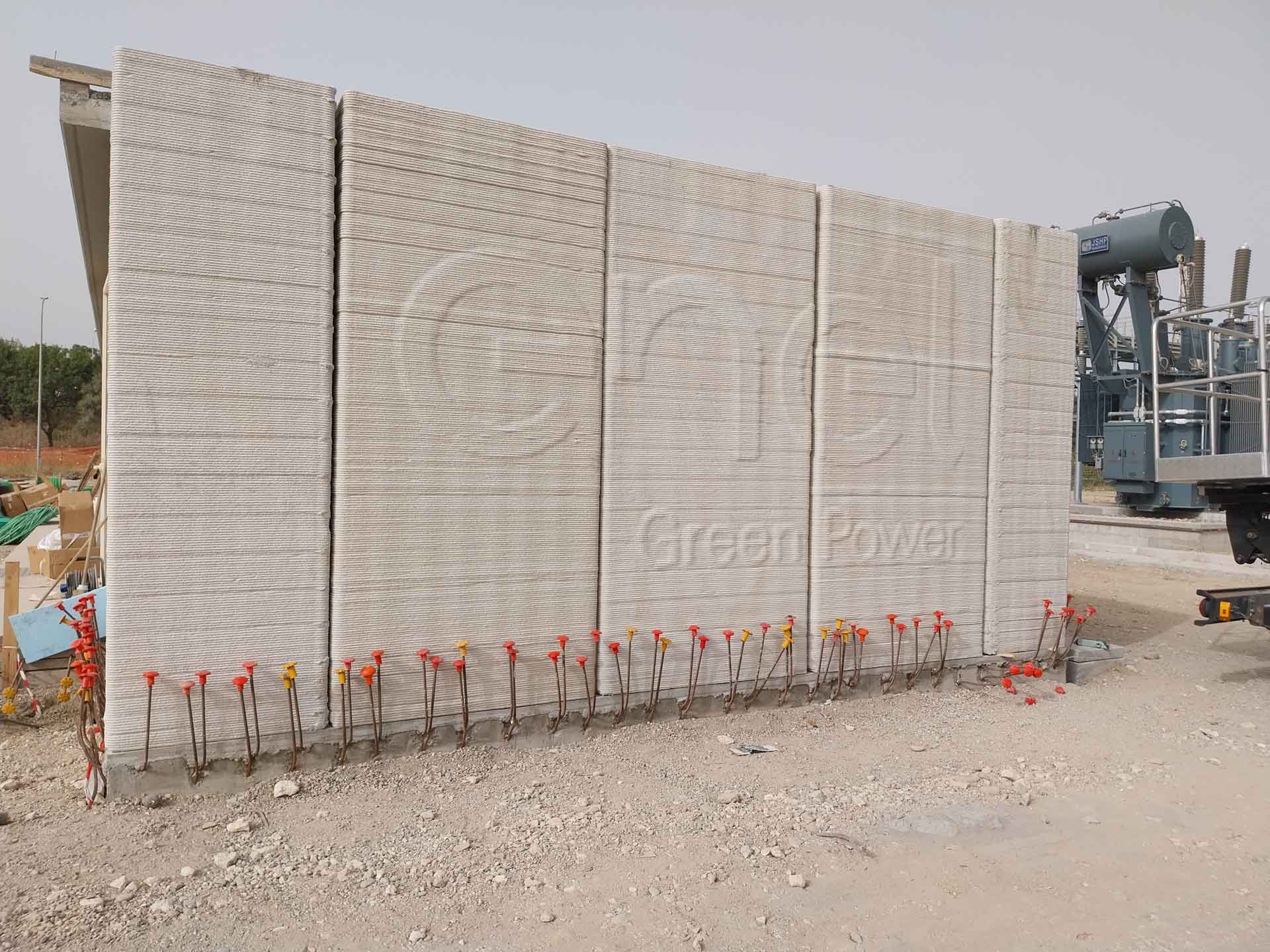 A large vertical 3D-printed concrete wall with a wavy texture and the imprinted words "Enel Green Power", with steel reinforcing bars capped with orange safety covers in front, at a construction site.