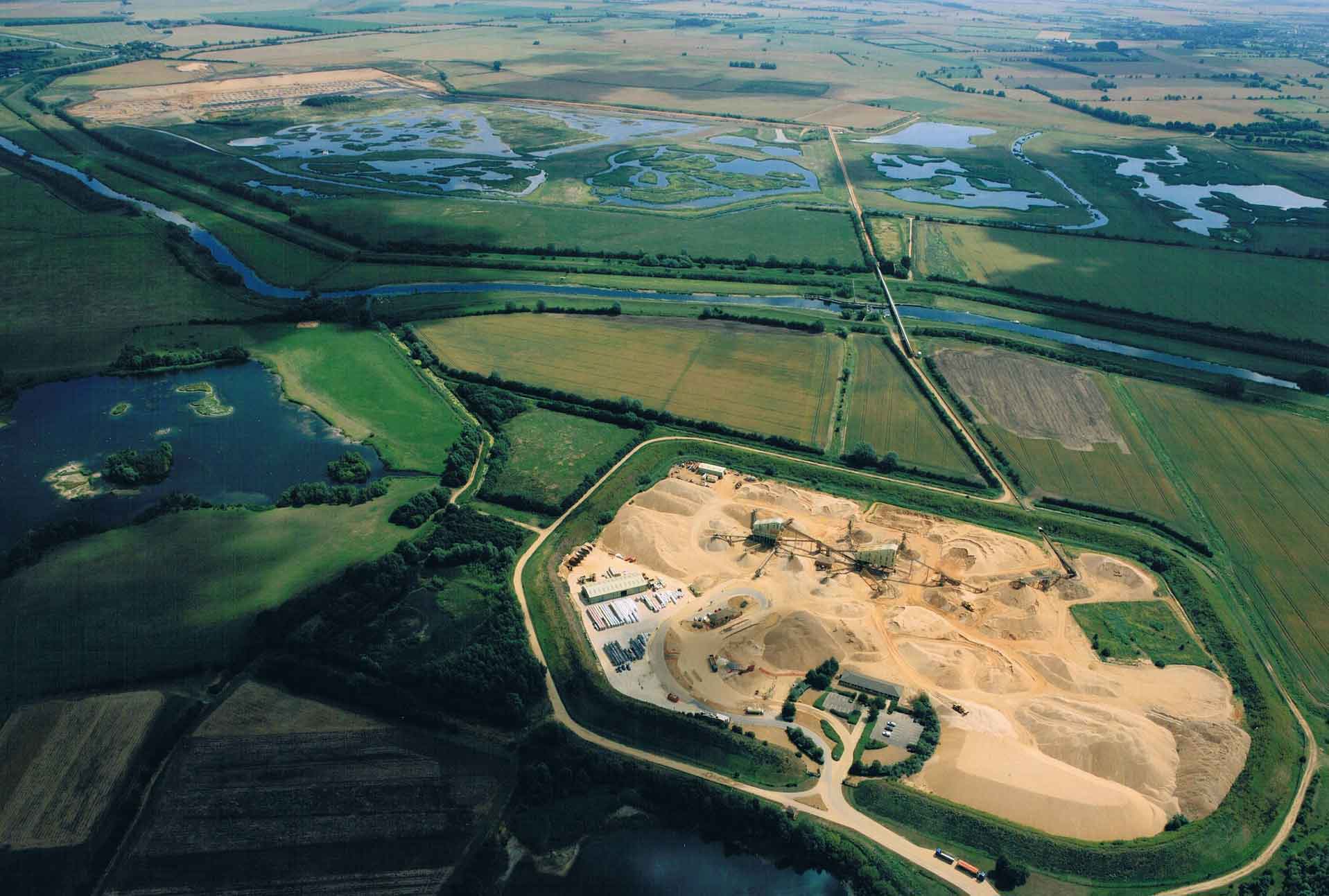 Aerial view of a wetland with a meandering river and a quarry with machinery at work in the lower right, surrounded by lush green fields.