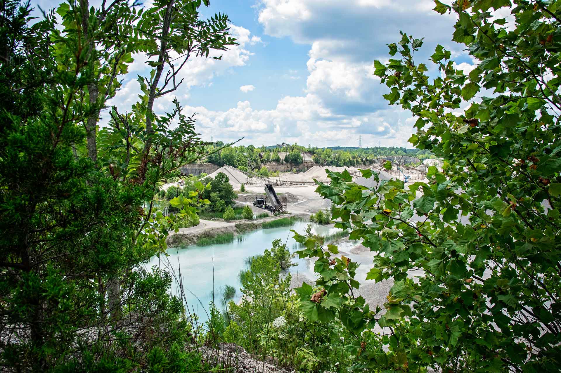 A scenic view of a quarry through foliage with a turquoise lake surrounded by gravel piles and industrial equipment.