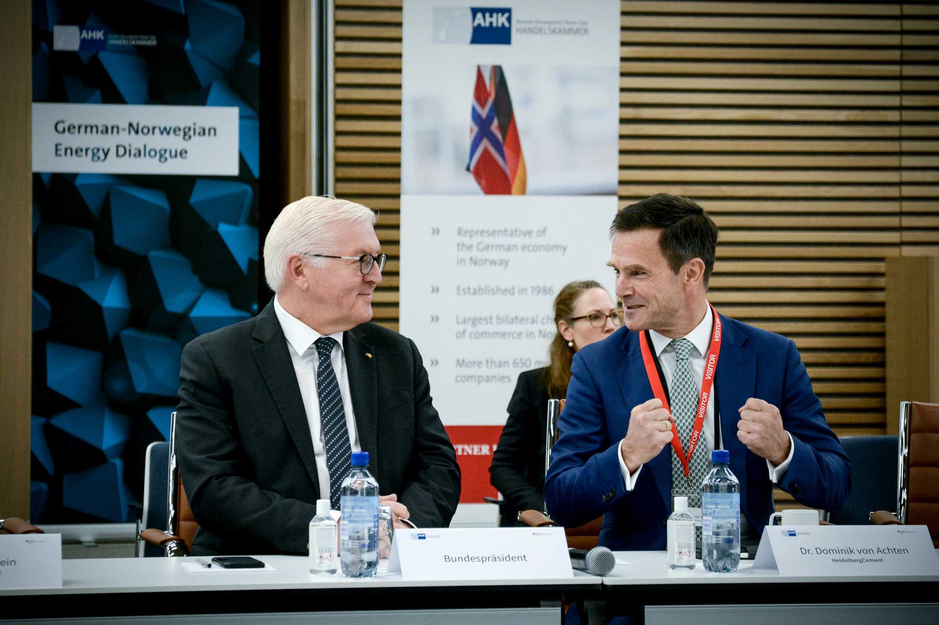 Frank-Walter Steinmeier and Dominik von Achten are seated at a table with microphones, engaged in a formal discussion. A partially visible sign in the background describes the meeting setting.