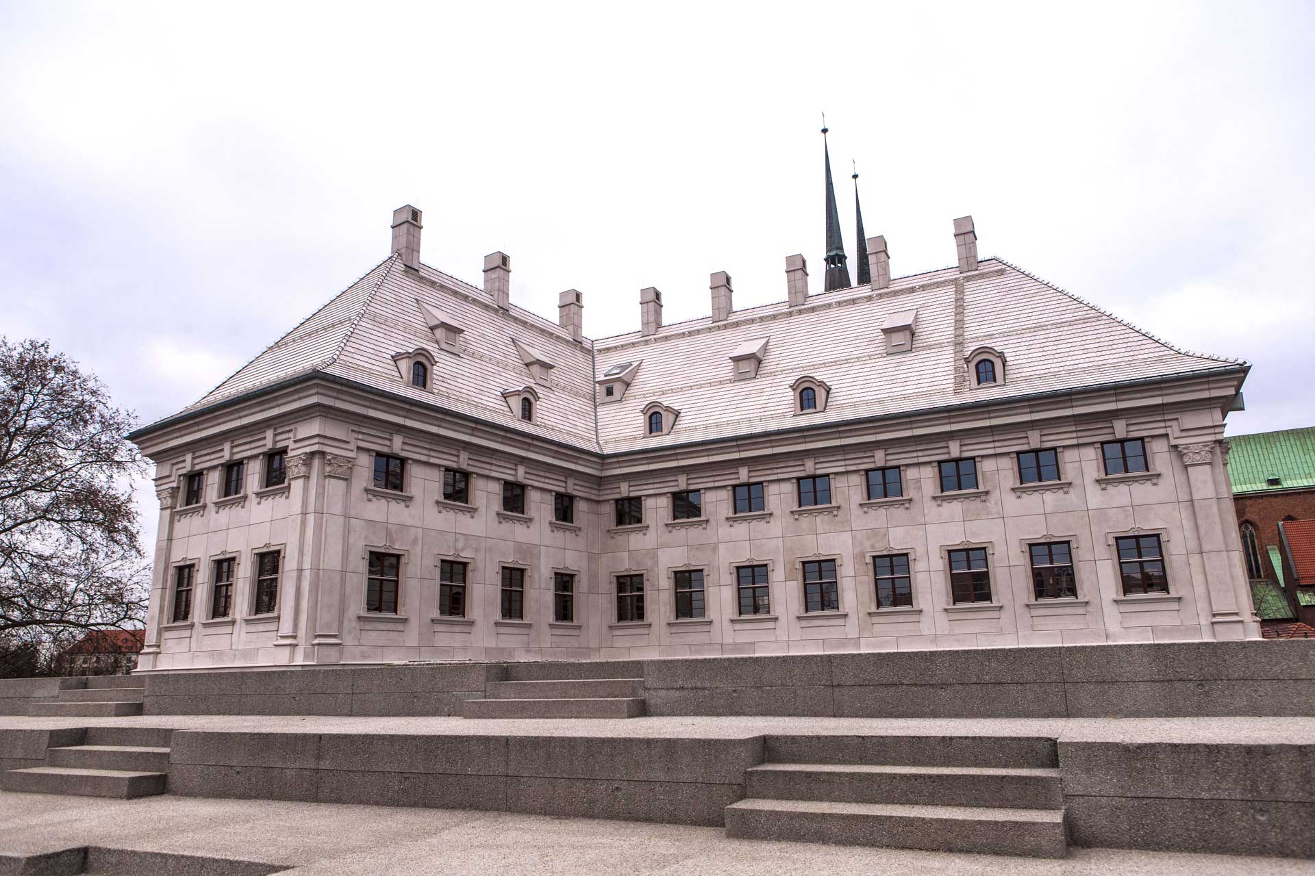 A grand, white stone building with a tiled roof and multiple windows. The building is set on a raised platform with steps leading up to it.