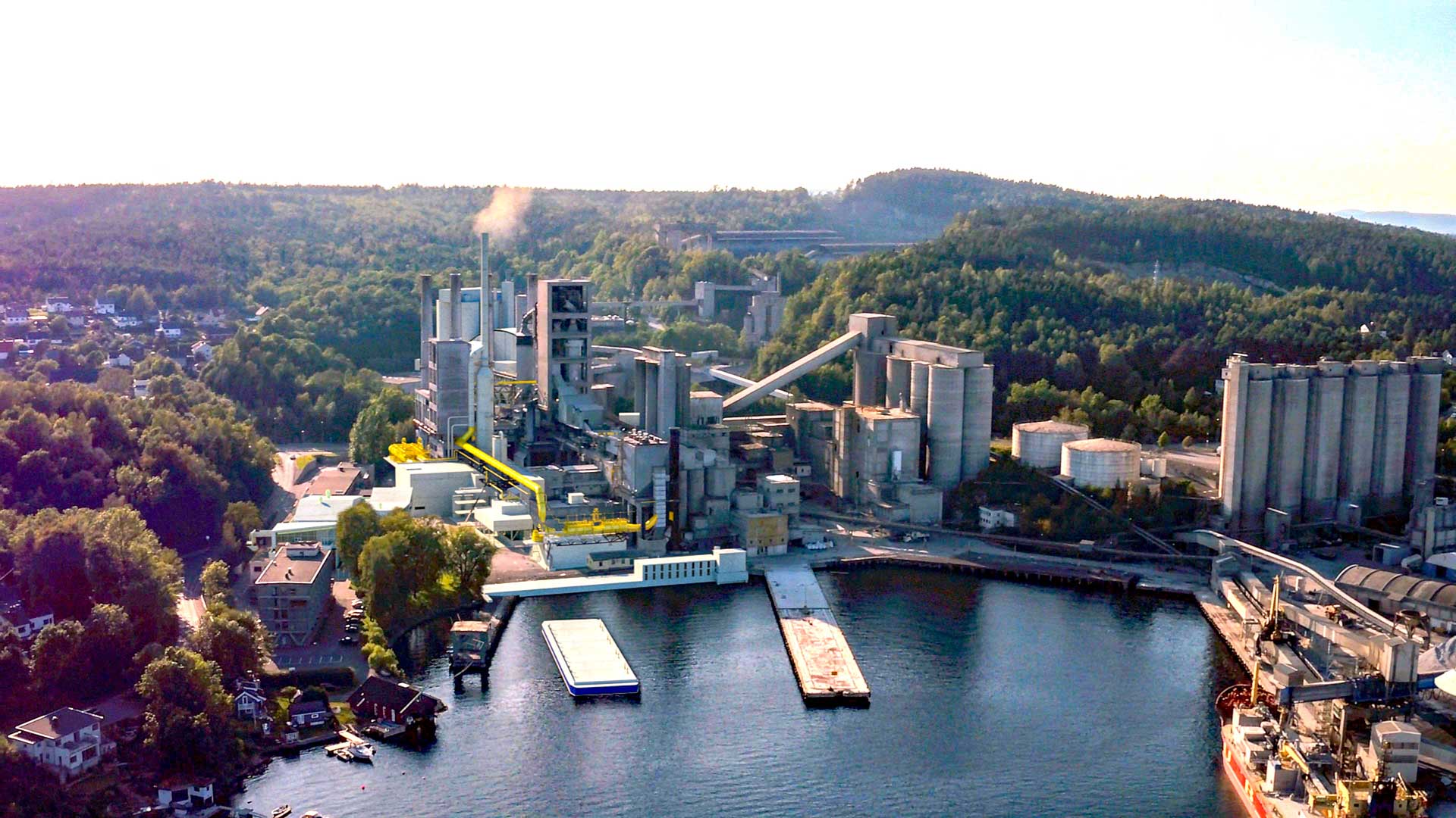 An aerial view of a cement plant with large structures, including silos, towers, and buildings connected by a network of pipes. A ship is docked at an adjacent body of water, surrounded by dense greenery.