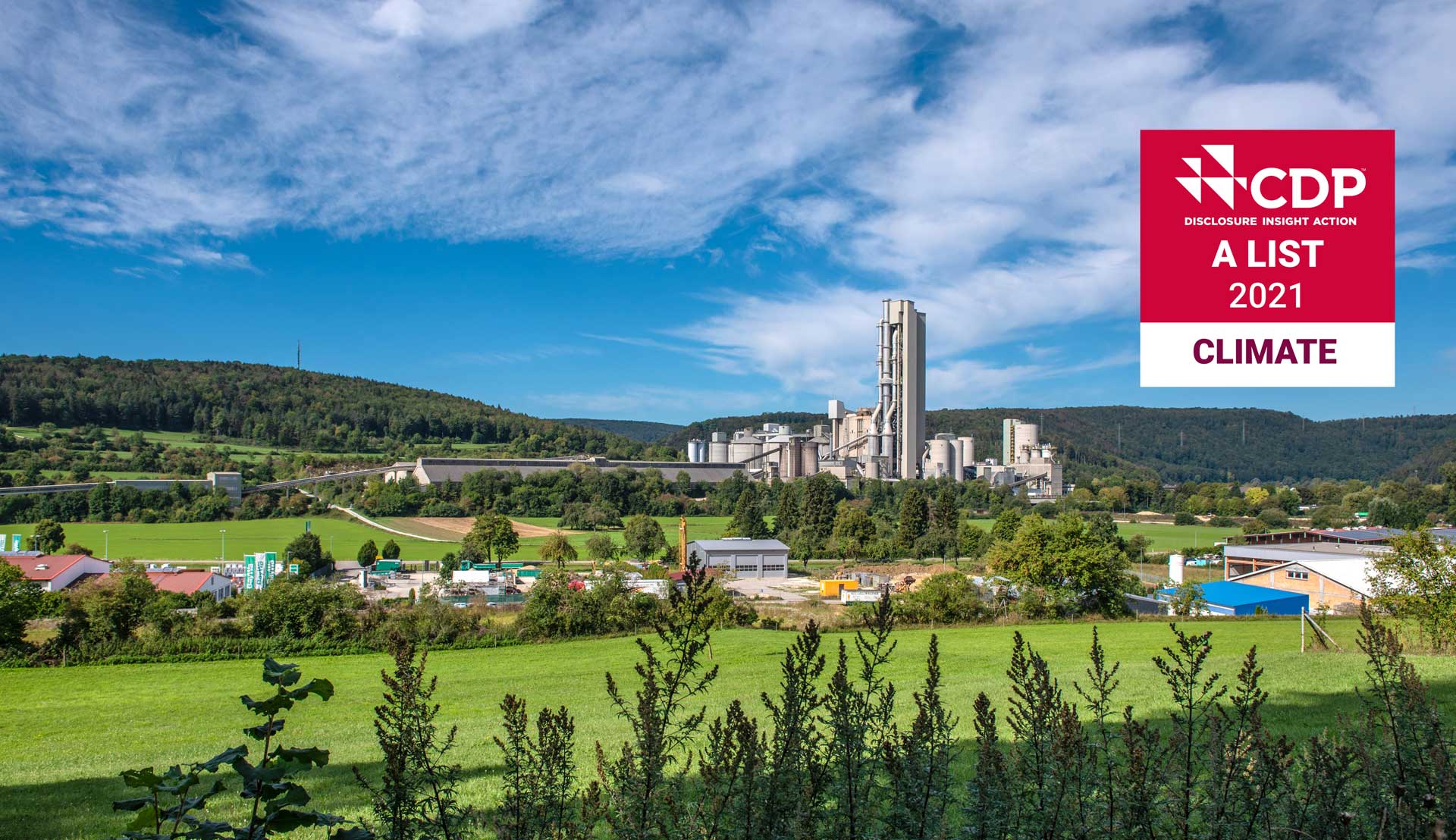 A cement plant nestled in a rural landscape, surrounded by green fields and hills. The plant has a large, white silo and several other structures. The CDP logo is visible in the bottom right corner, indicating that the plant is part of the CDP Climate Change Program.