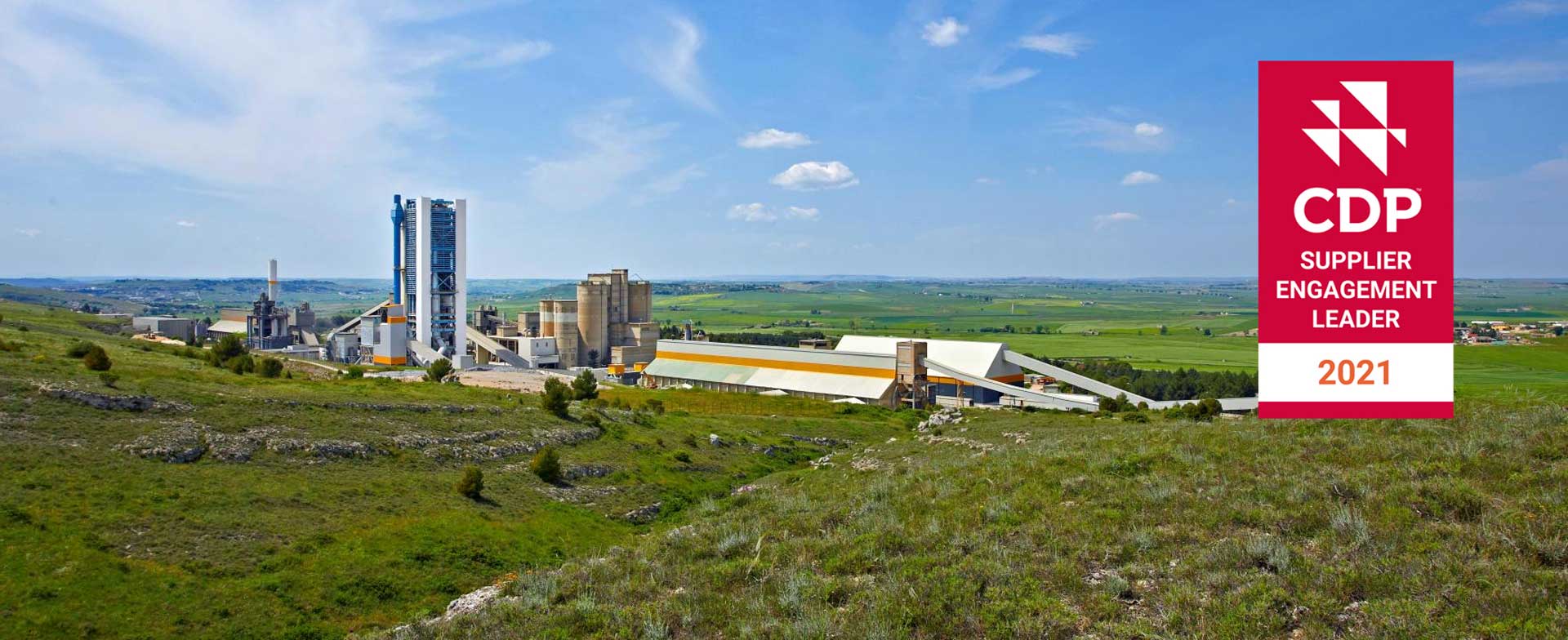 A cement plant nestled in a rural landscape. The plant consists of several large industrial buildings and structures surrounded by green fields and hills. A clear blue sky with white clouds is visible above. In the foreground, there is a red banner with the CDP logo and the text "SUPPLIER ENGAGEMENT LEADER 2021."