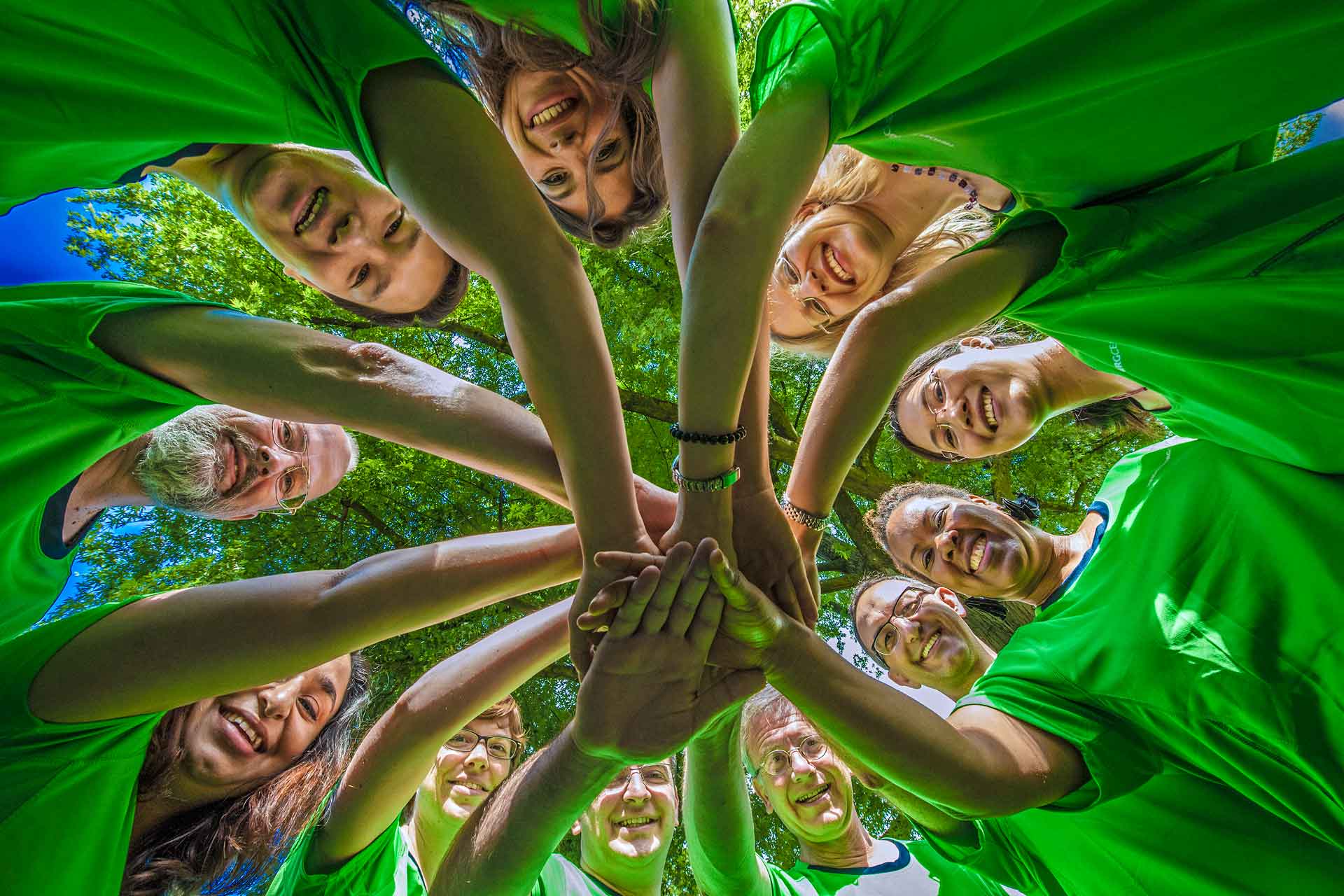 View from below: People stand in a circle, their hands meet in the centre. They are all wearing green T-shirts.