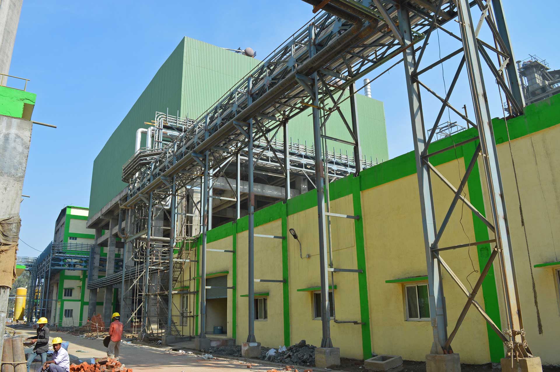 Green and yellow cement plant with metal pipelines on the right and two workers with helmets below under a clear blue sky.