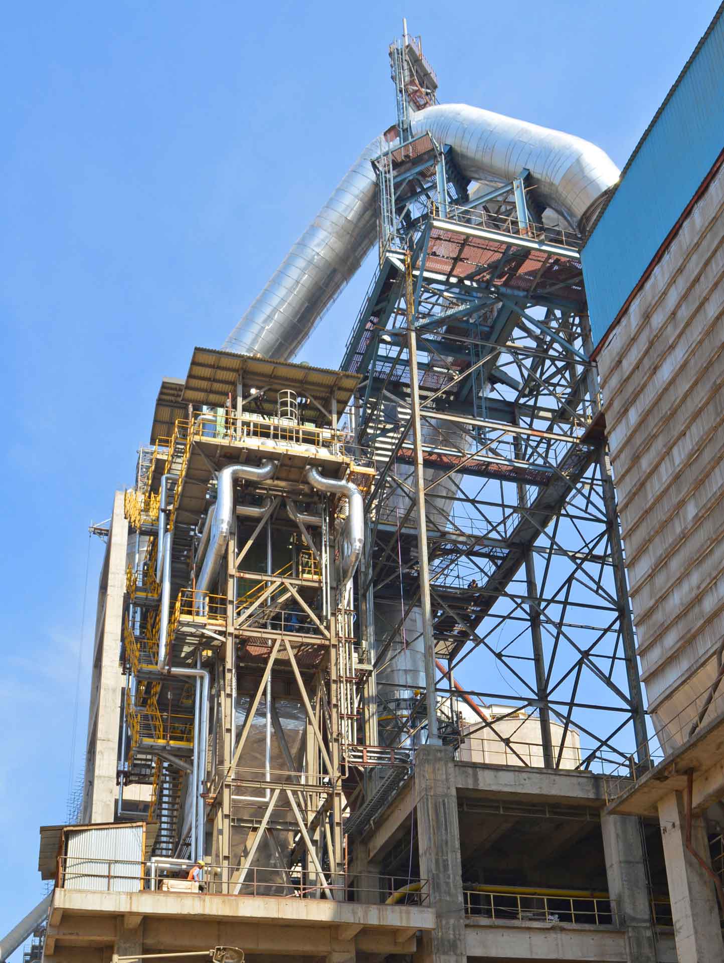 A large industrial facility with tall metal structures, pipes, and machinery. The sky is blue and clear.