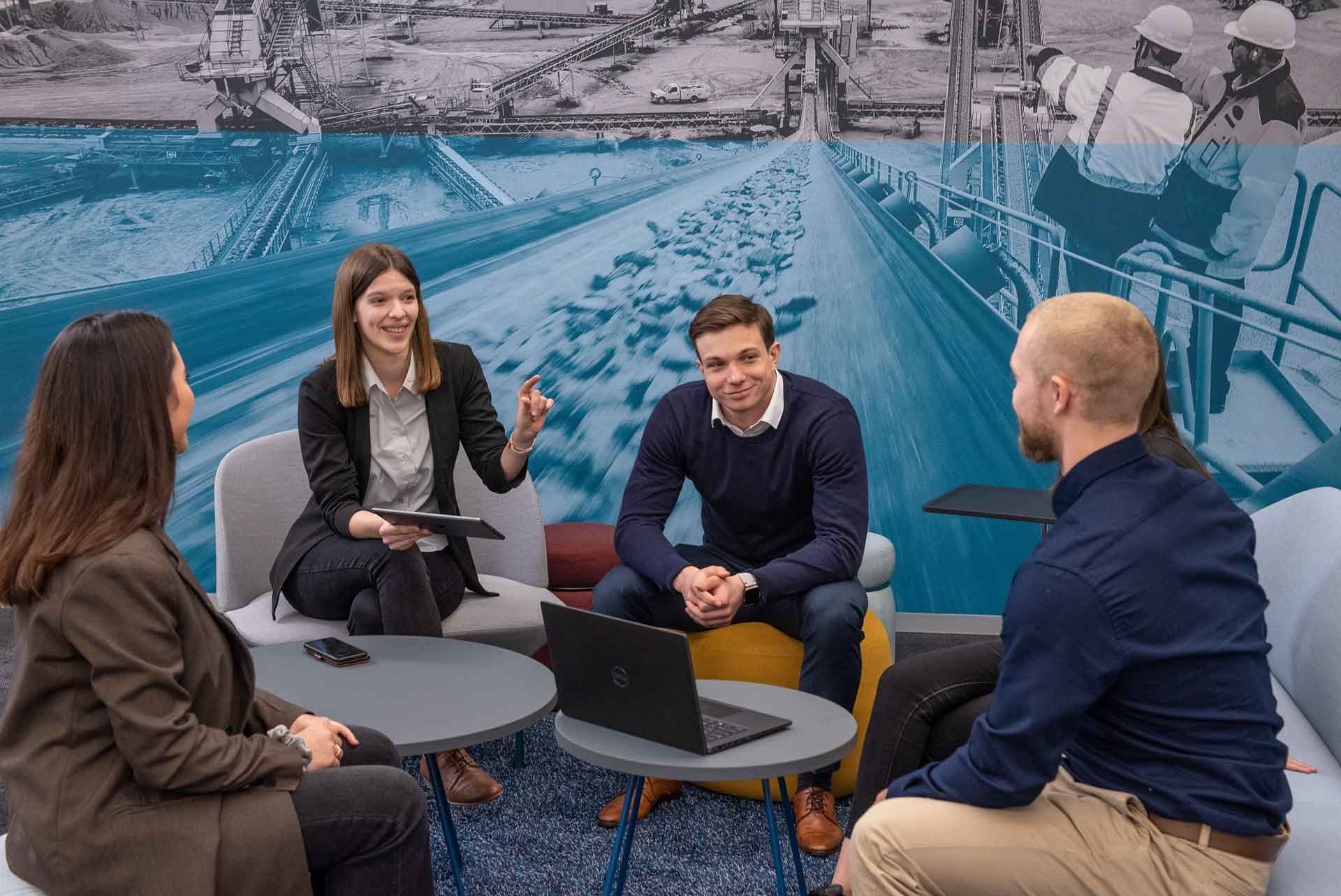 Vier junge Menschen im Business-Casual-Look sitzen um einen niedrigen runden Tisch herum, im Hintergrund das Bild eines blau eingefärbten Förderbandes