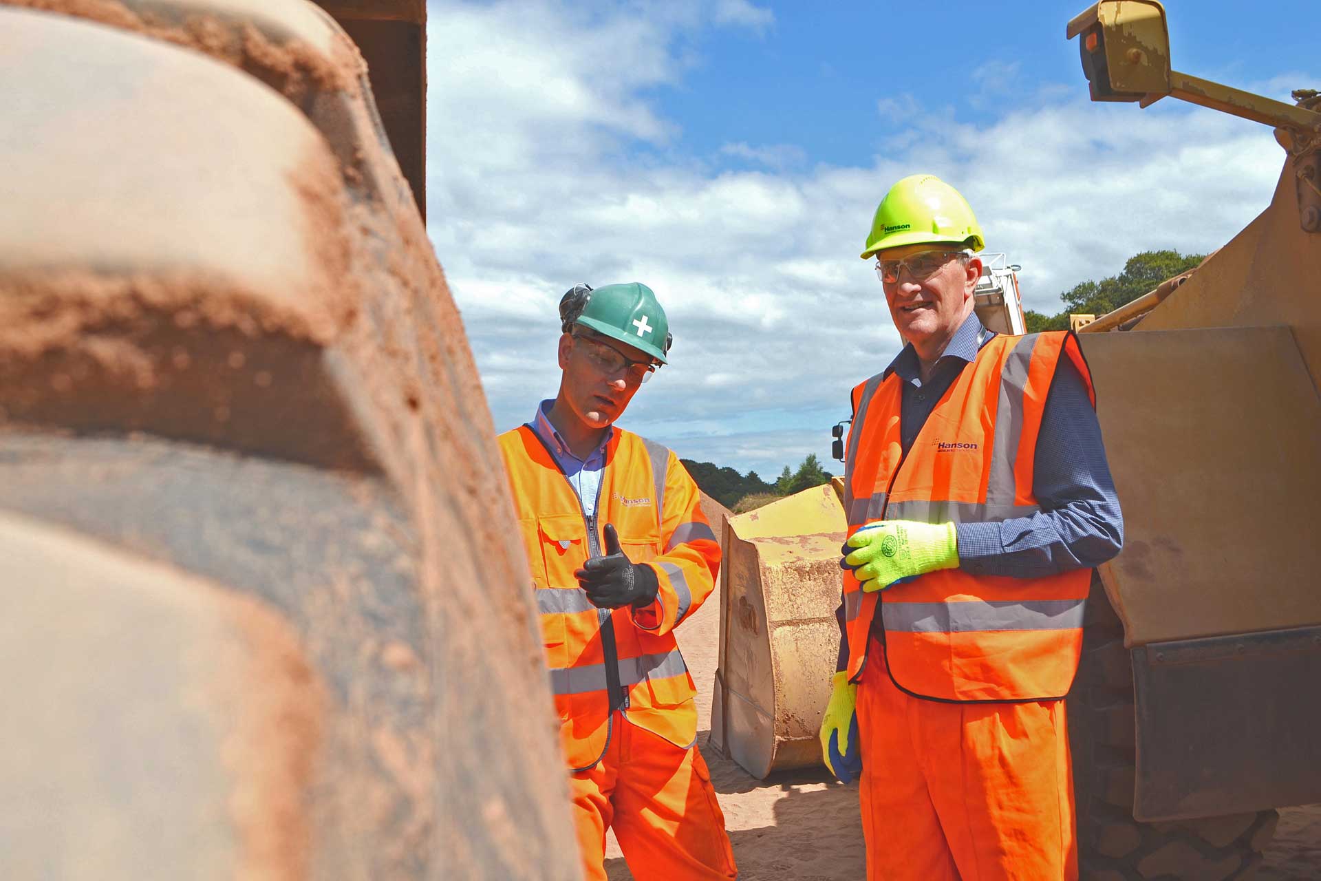 Two individuals in safety clothing and hard hats at a construction site, one holding a clipboard, engaged in discussion. A blurred wall is visible in the foreground.