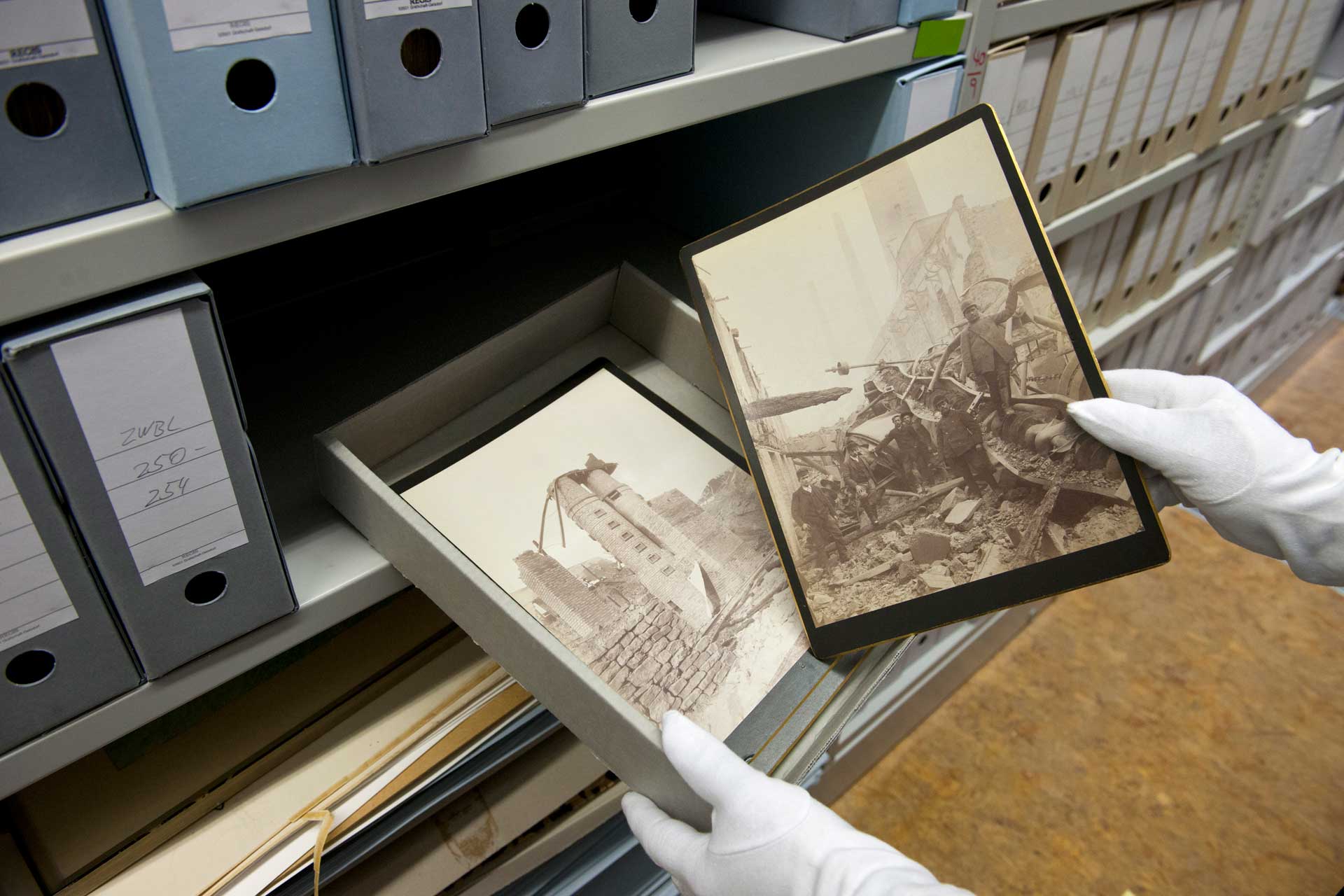 Hände mit Handschuhen halten eine Schachtel mit historischen Fotos, eine Hand hält dabei eines der Fotos