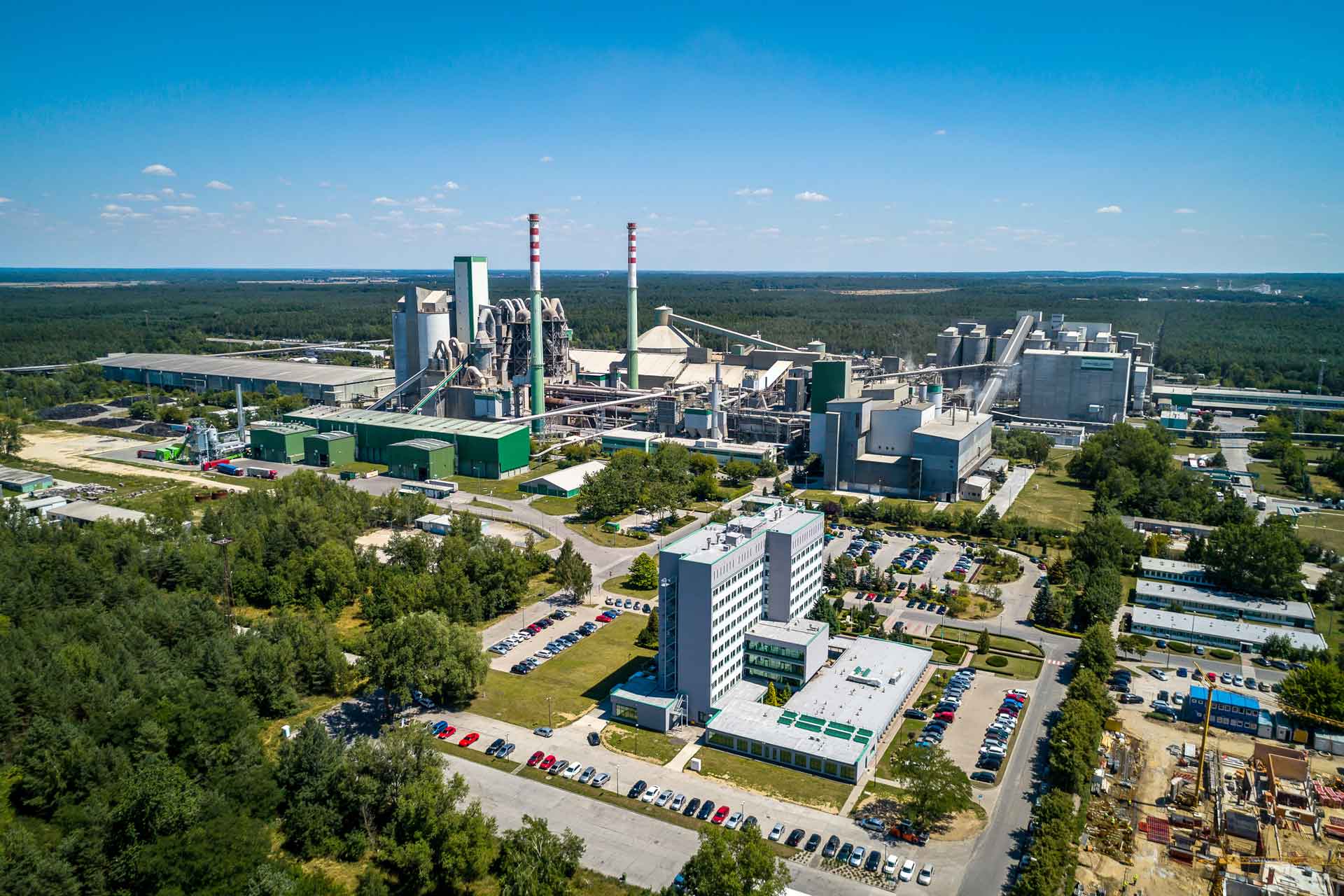 An aerial view of a large cement plant with multiple buildings, silos, and smokestacks. The plant is surrounded by trees and a parking lot.