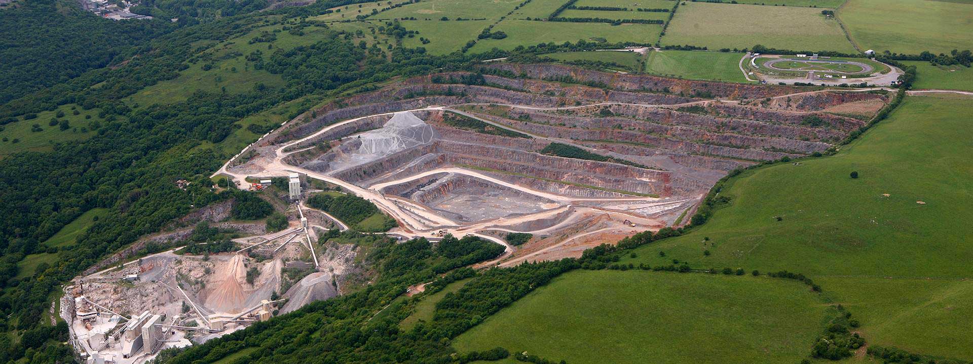 Aerial view of an expansive quarry with terraced excavation levels, surrounded by lush greenery and connected by a winding road.