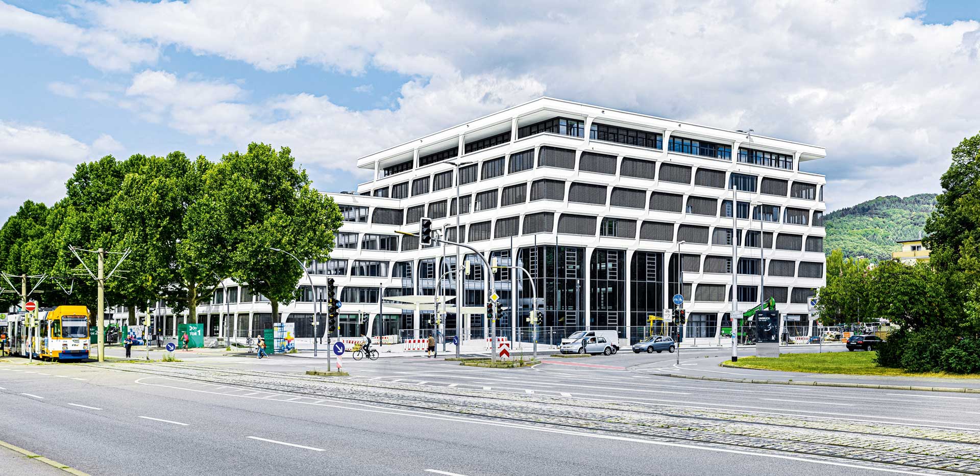 A modern office building with a glass facade and a central courtyard. There are trees and a tram line in front of the building.