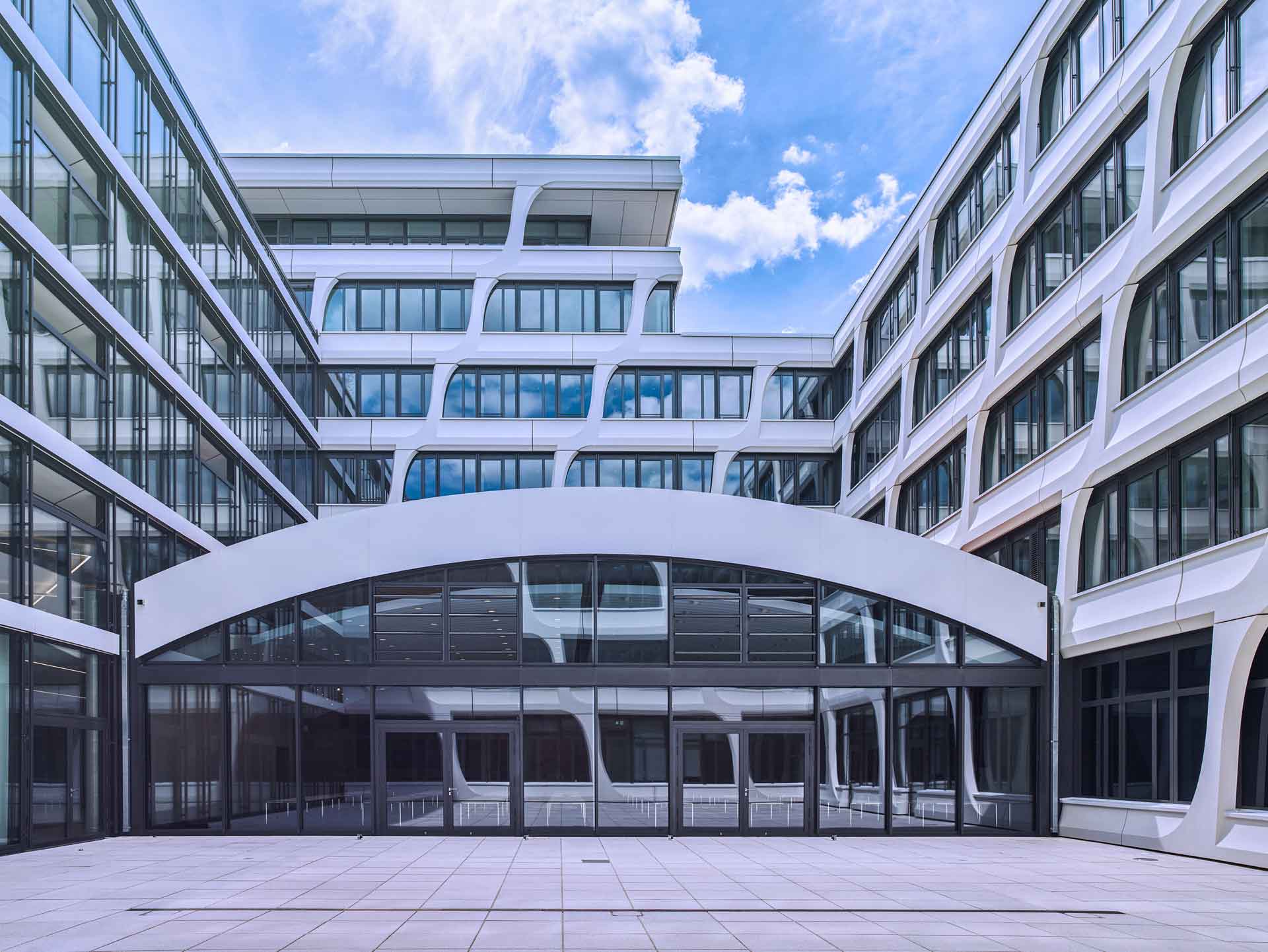 A modern, white office building with a curved glass atrium and a central courtyard. The building is surrounded by other office buildings and has a blue sky in the background.