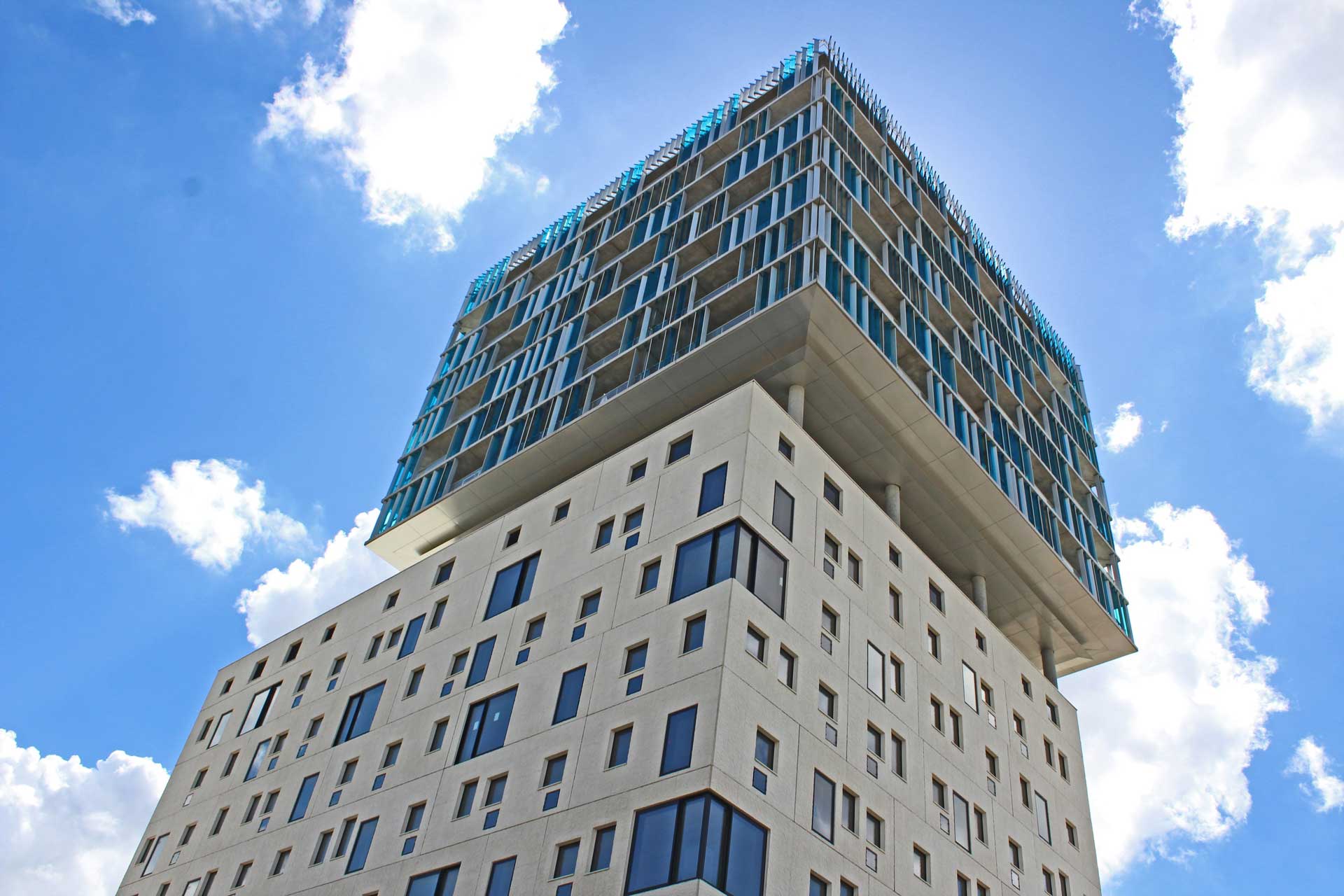 A modern high-rise building with a unique design, featuring a larger upper section that appears to float above the narrower lower section against a background of blue sky and clouds. 