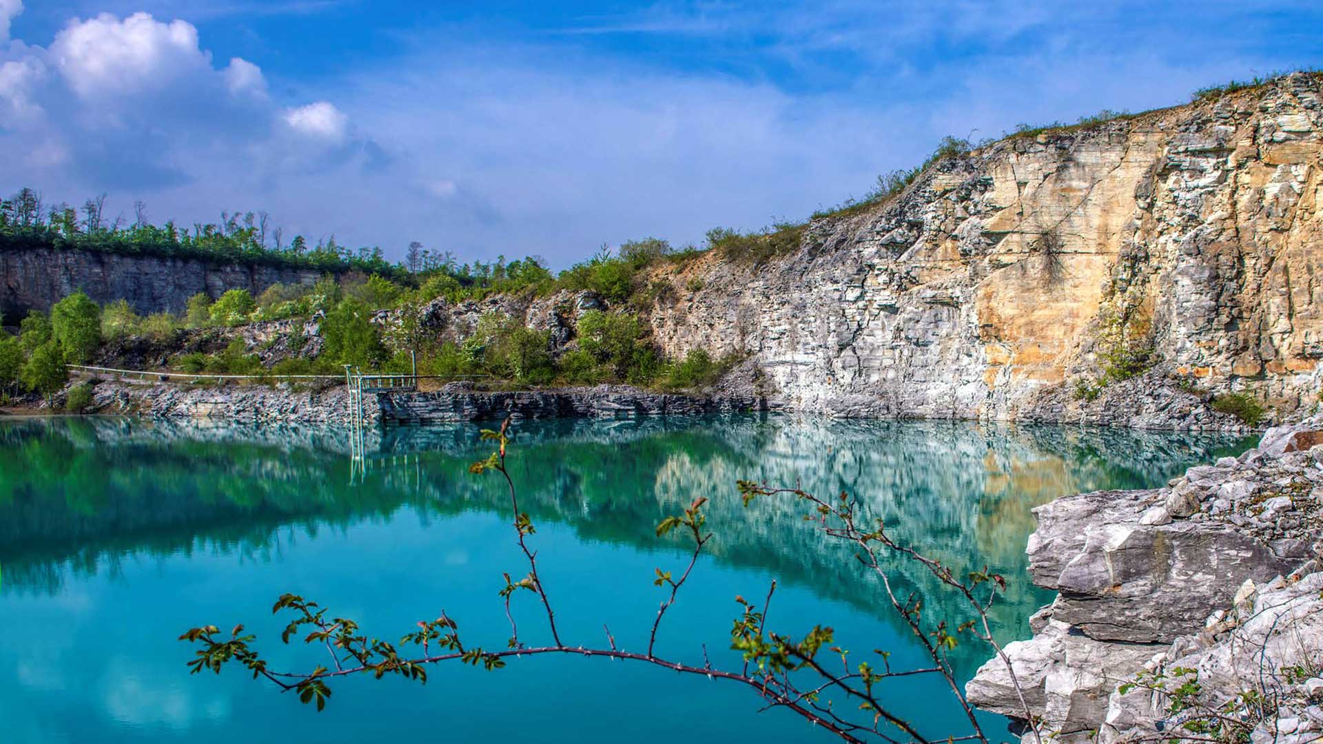 Malerischer Blick auf einen klaren blauen See, umgeben von Feldwänden mit üppiger grüner Vegetation unter einem teilweise bewölkten Himmel.