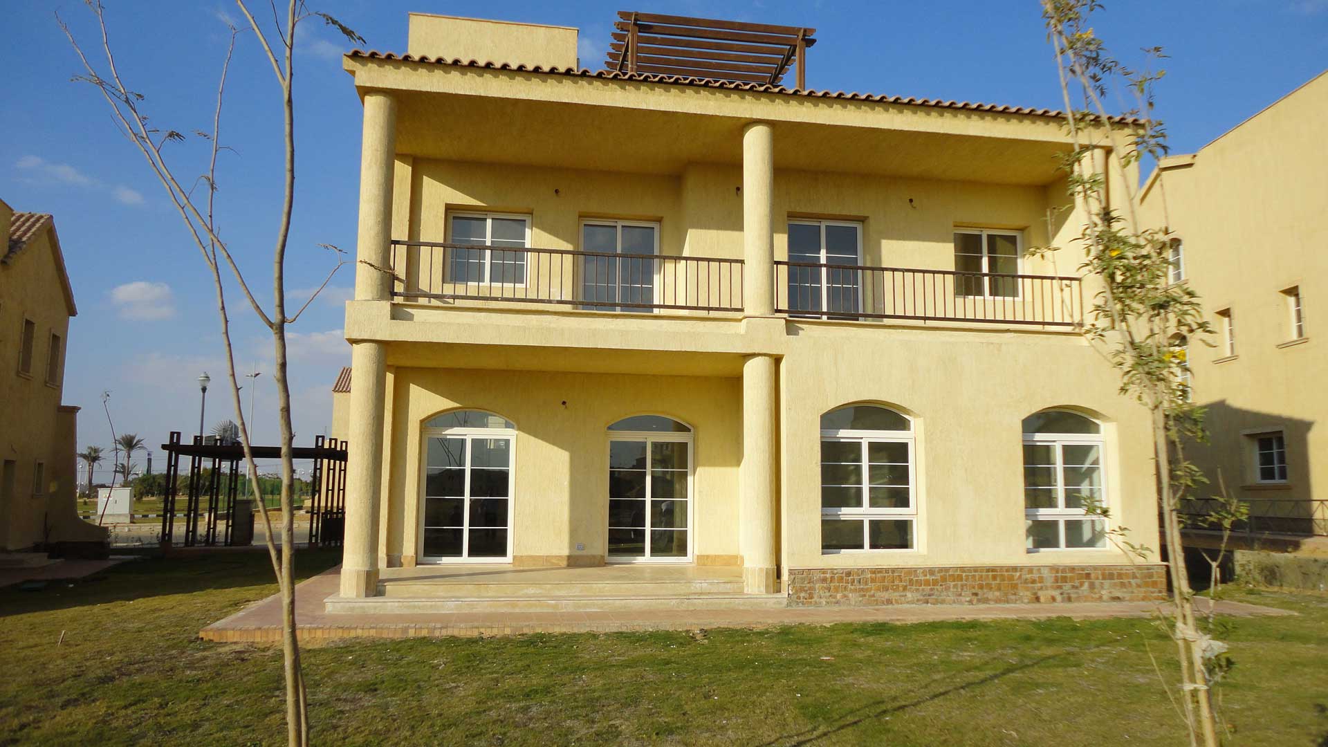 Eine zweistöckige Villa mit einer gelben Fassade und einem Terrakotta-Dach. Die Villa hat einen Balkon im zweiten Stock und eine Terrasse im Erdgeschoss. Im Hintergrund befindet sich ein Garten mit Palmen.