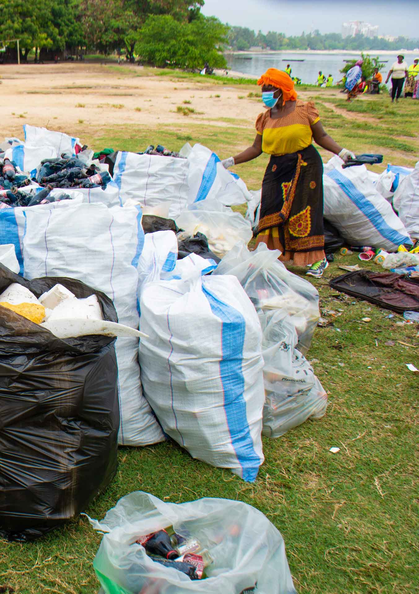 Woman in face mask standing among full rubbish bags, with trees and water in the background.