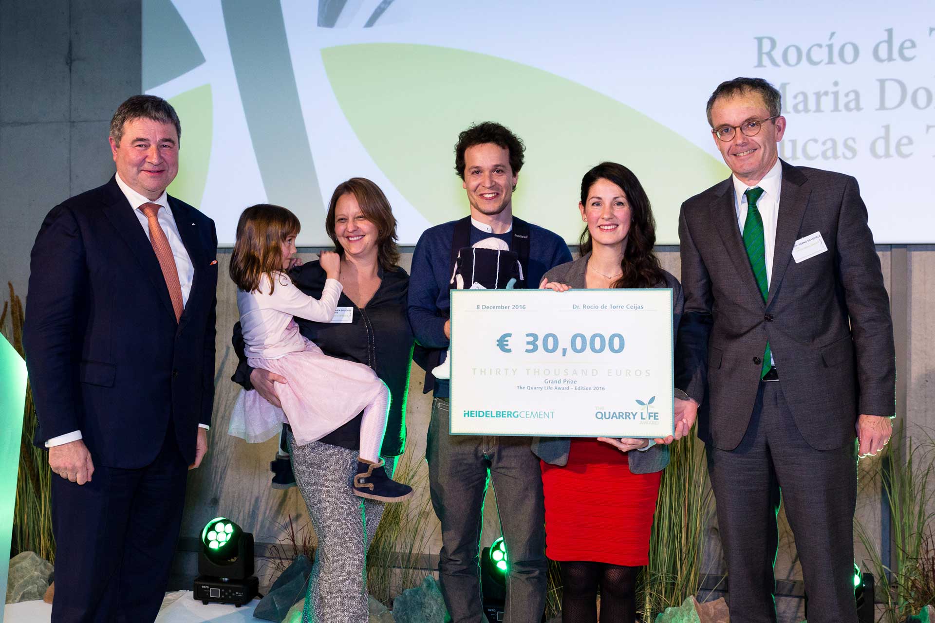 Seven people on a stage, one of them is a child in arms, one is a baby in a carrier. One person is holding a symbolic cheque with the imprint €30,000, HeidelbergCement and Quarry Life Award.