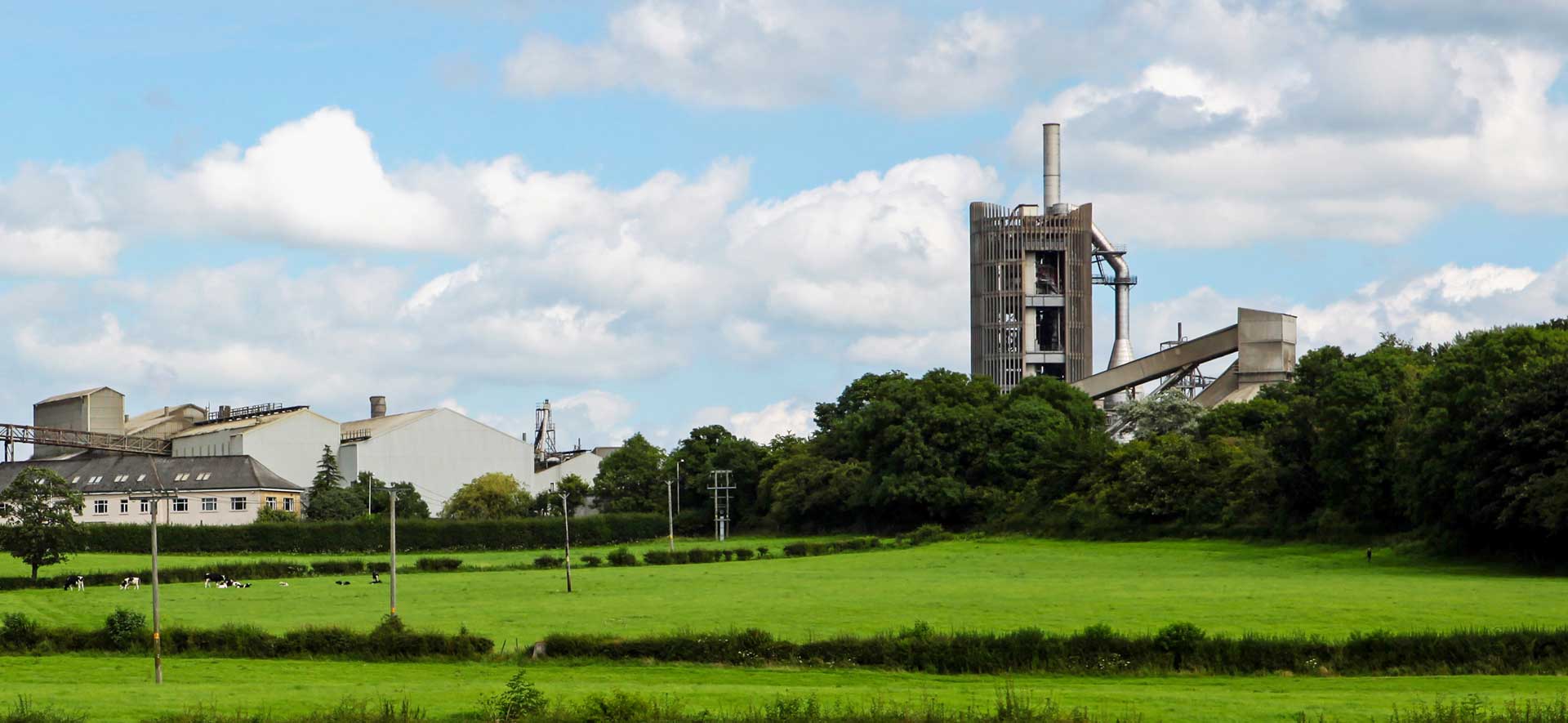 A cement plant with a large chimney and several buildings next to a green field under a partly cloudy sky.