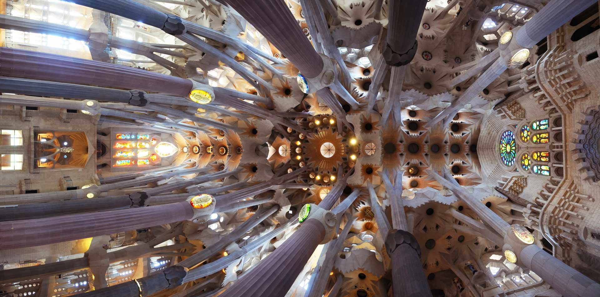 The interior of the Sagrada Família Basilica in Barcelona, designed by Antoni Gaudí, features a ceiling with intricate geometric patterns and a play of light and shadow created by columns and stained glass windows.