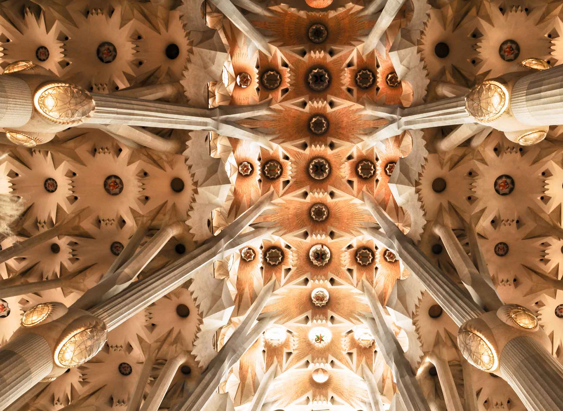 The image displays the complex ceiling architecture of a cathedral with detailed patterns and multiple adorned columns tapering towards the center. The design features a symmetrical arrangement of star-shaped elements and circular motifs, enhanced by a warm color palette that underscores the visual intricacy.