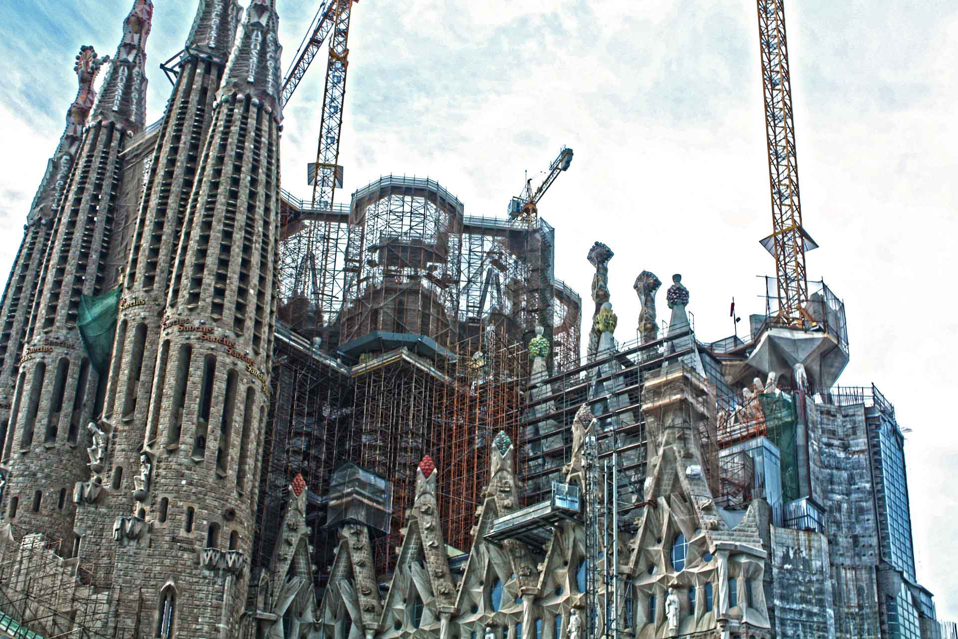 A majestic cathedral with complex facades and towers is under construction, surrounded by numerous cranes and scaffolding.