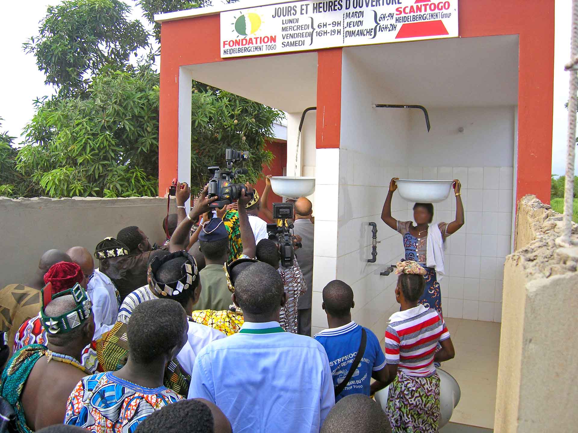 A group of people at a water pump station with a sign in French indicating the foundation’s involvement and operating hours.