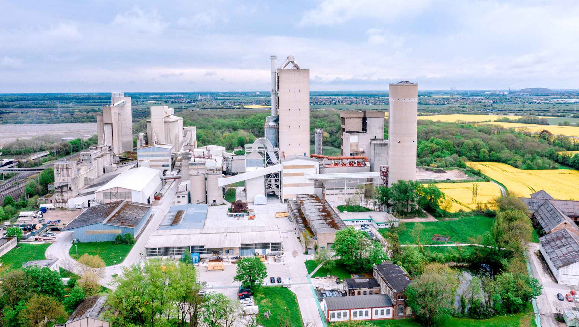 An aerial view of a large cement plant with multiple buildings and silos, surrounded by green fields and a nearby town.