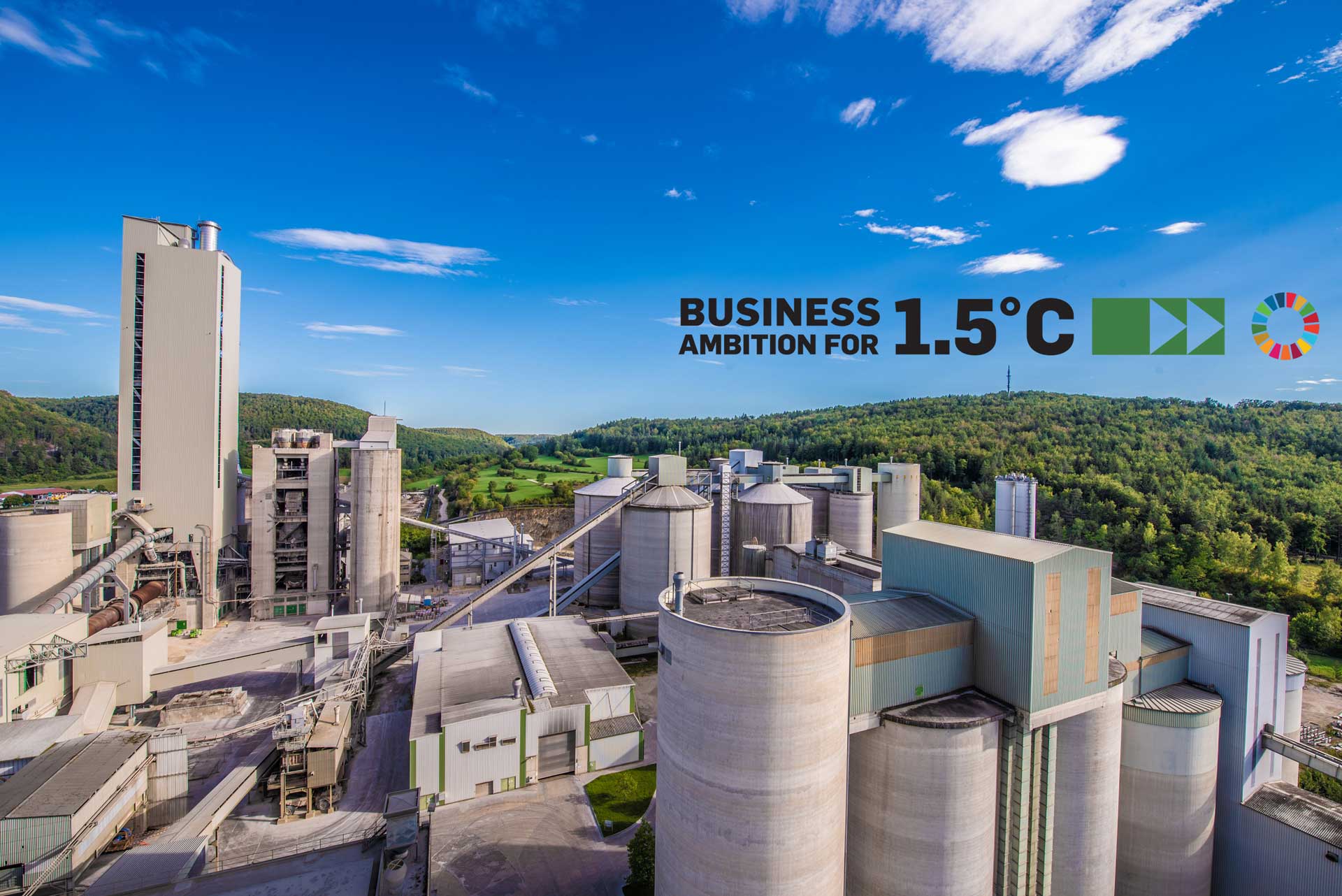 Ein Zementwerk mit mehreren Silos und Gebäuden, umgeben von grünen Hügeln und einem blauen Himmel. Die Wörter "BUSINESS AMBITION FOR 1.5°C" und das Logo der Vereinten Nationen sind in der oberen rechten Ecke sichtbar.