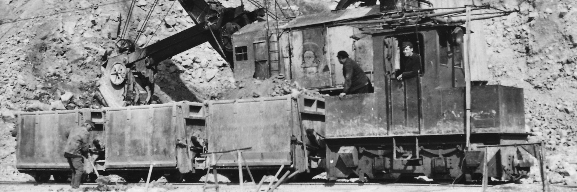 Historisches Foto mit einem Triebfahrzeug vor drei Loren, in dem Fahrzeug stehen zwei Arbeiter