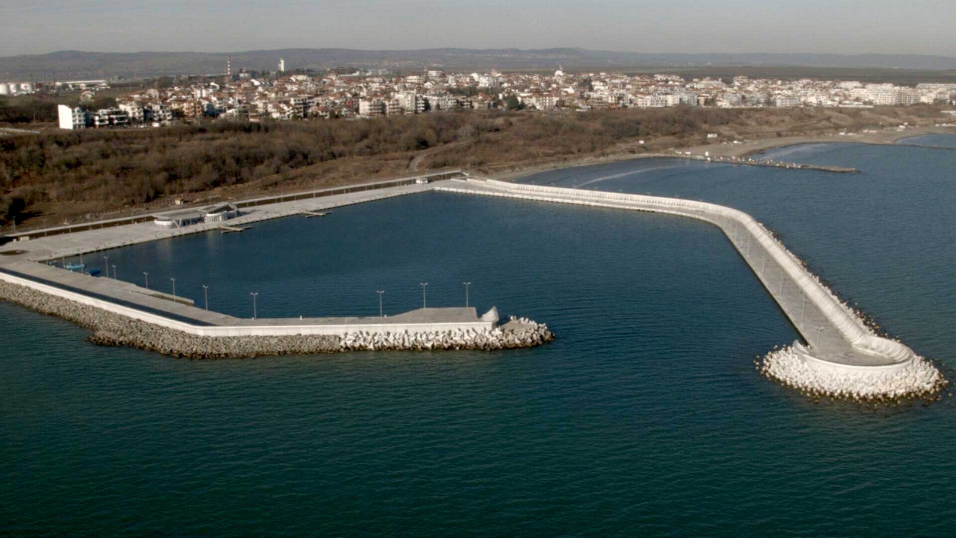 An aerial view of a man-made harbor with a breakwater extending into the sea. The harbor is surrounded by a coastal town and a large body of water.