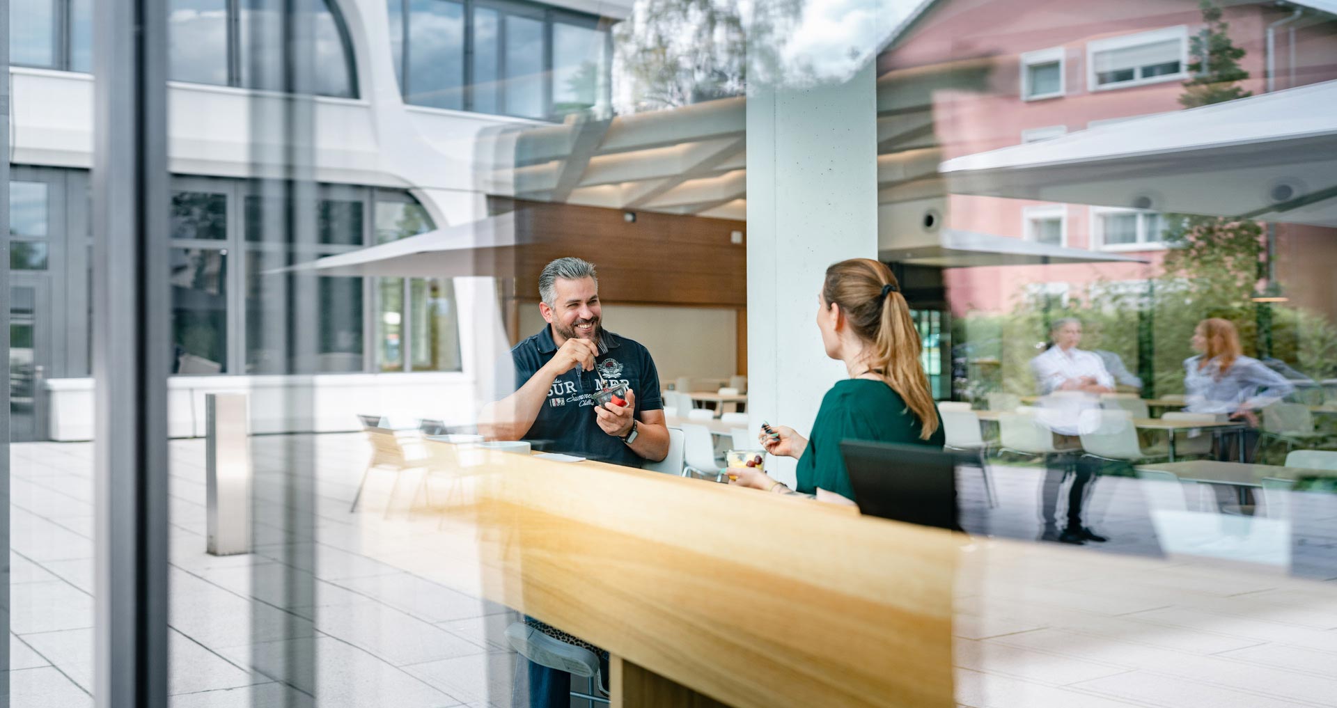 Zwei Personen sitzen an einem Tisch hinter einer teilweise reflektierenden Glasscheibe und essen etwas