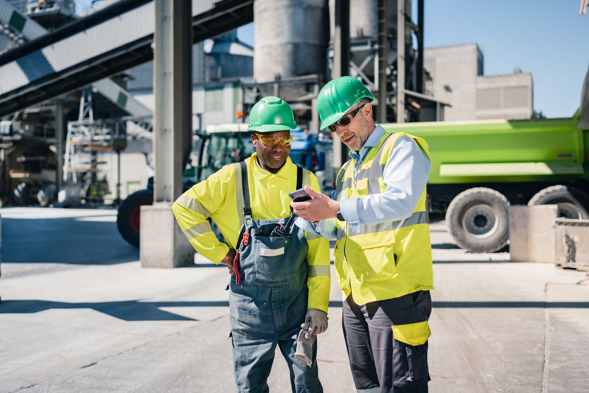 Two men with helmets standing in front of a cement plant, one person shows the other something on a smartphone.