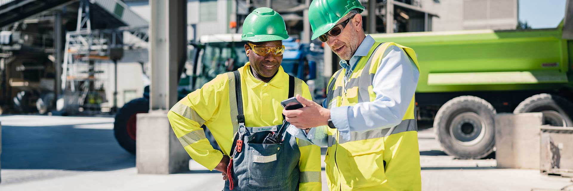 A construction worker in a yellow vest shows their colleague something on his mobile phone