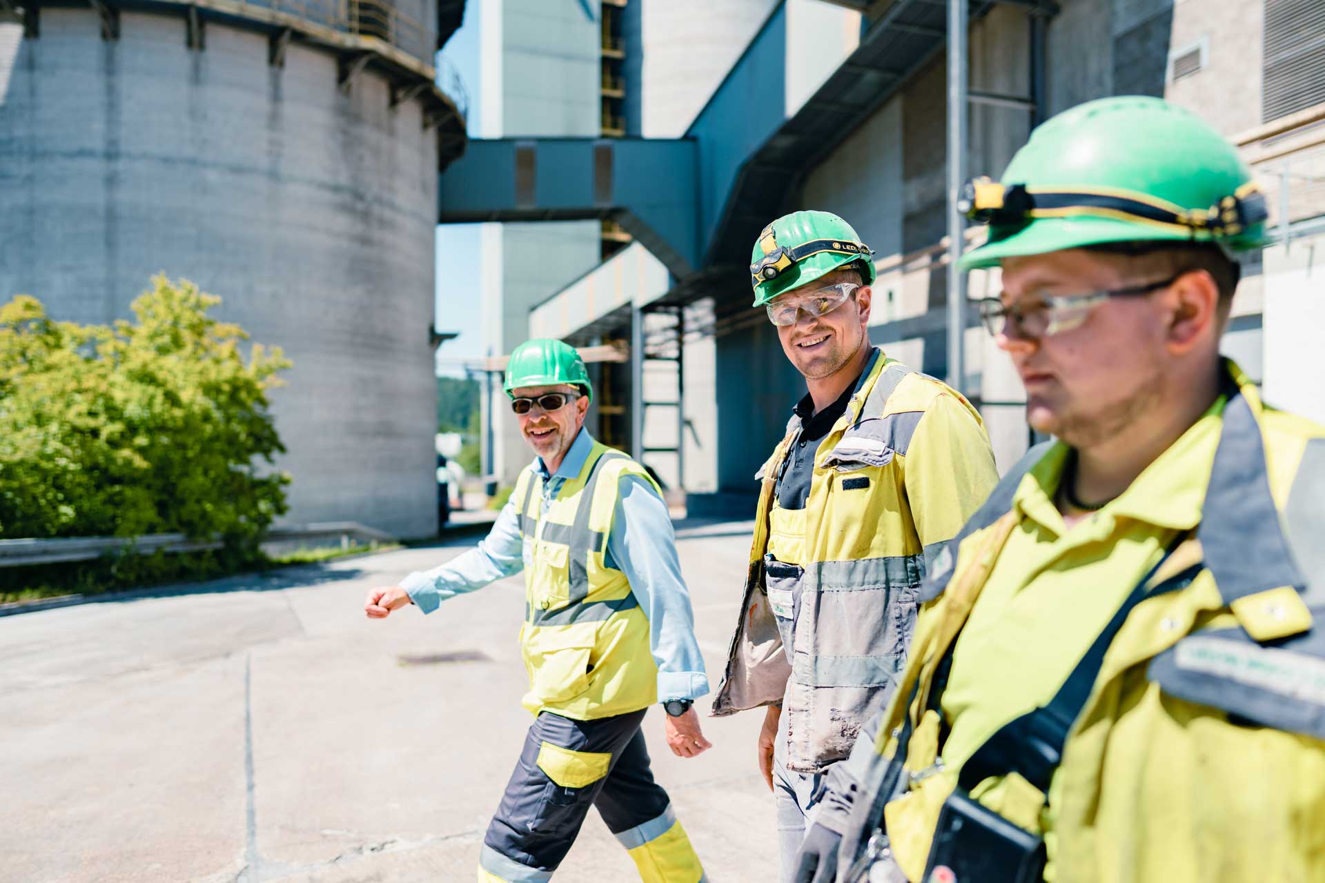 Drei Personen in Schutzkleidung laufen nebeneinander über ein Industriegelände mit verschiedenen Gebäuden