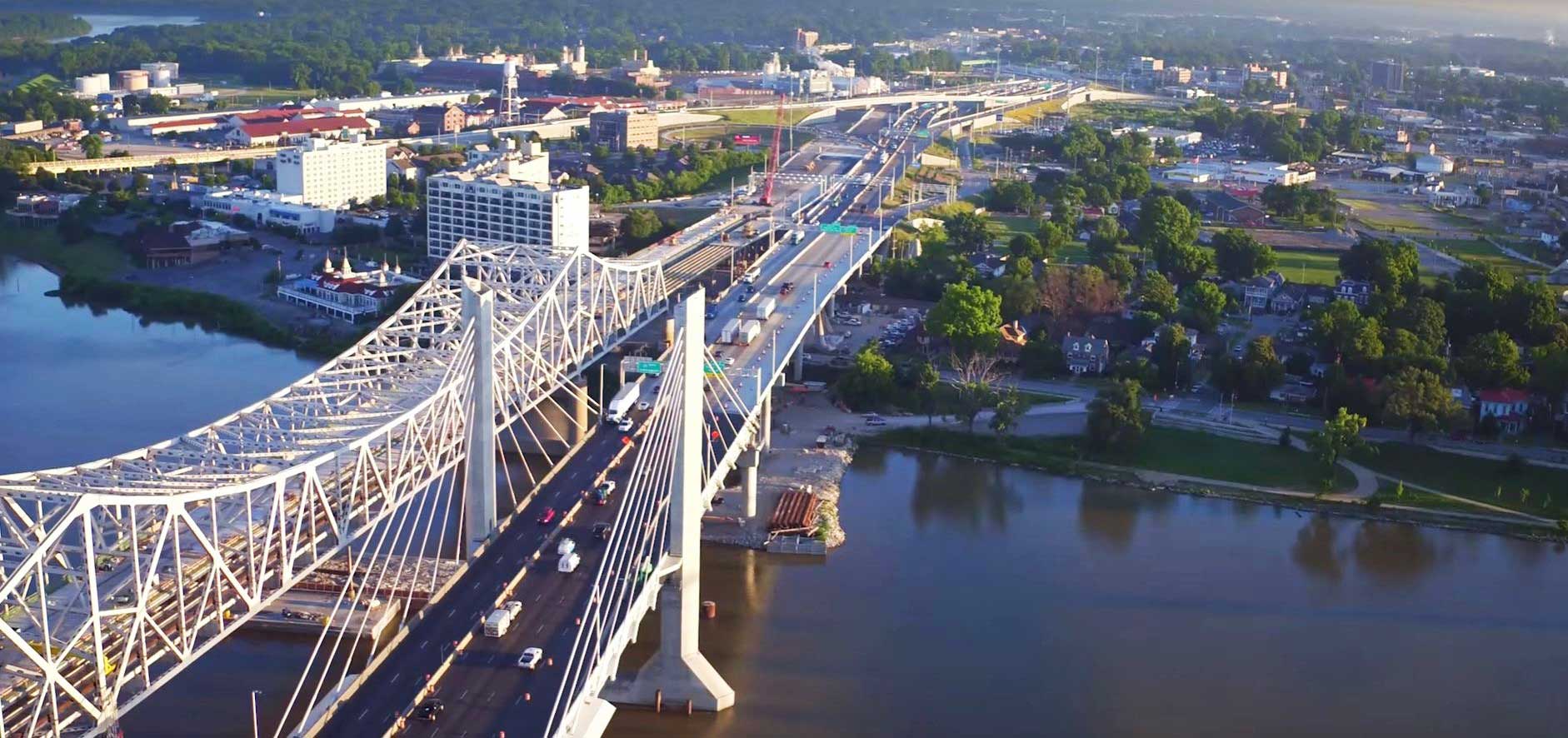 Aerial view of a large suspension bridge spanning a river, with a city skyline in the background.