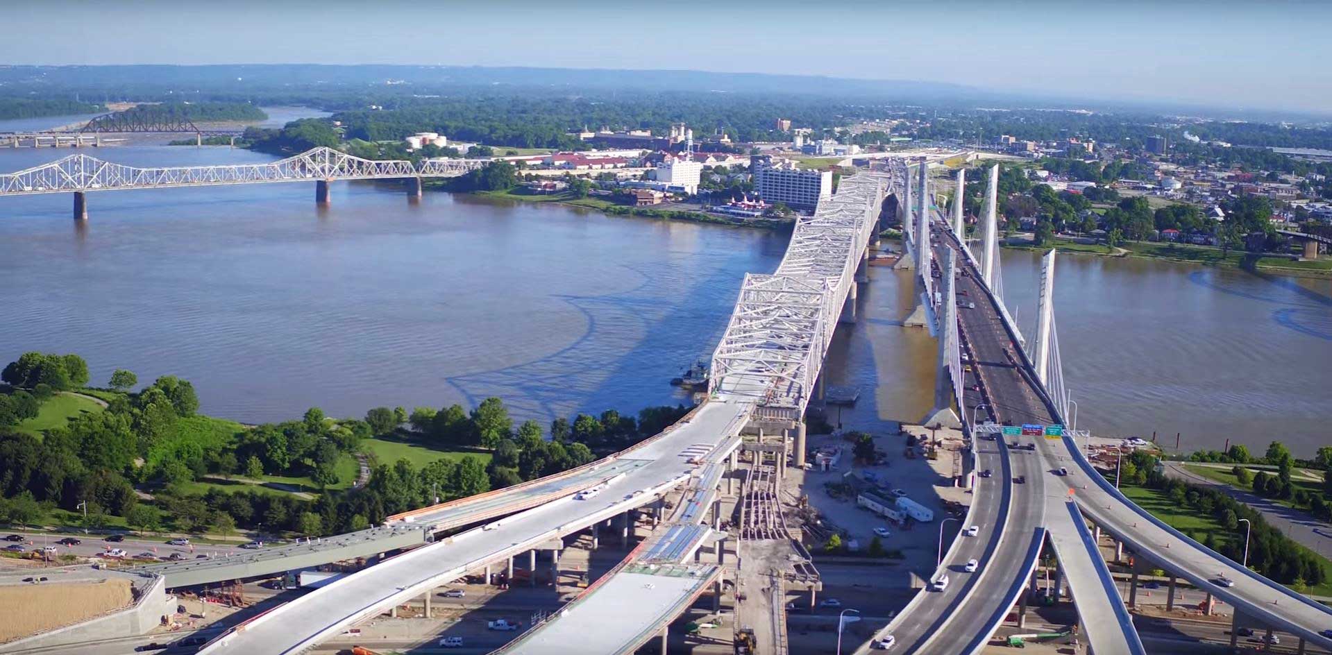 Aerial view of a large cable-stayed bridge under construction. The bridge spans a wide river, and there are several roads and interchanges visible around it.