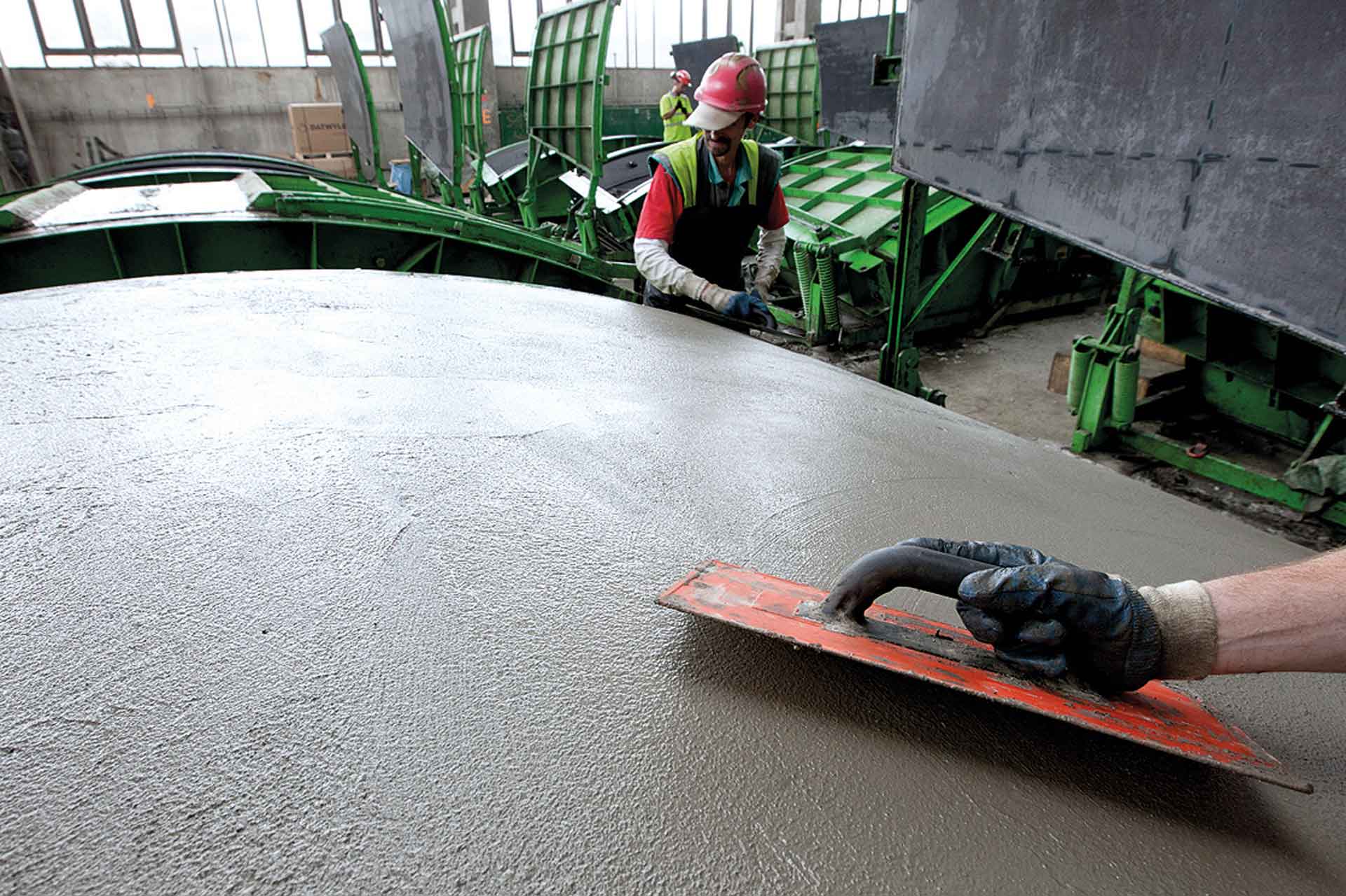 Zwei Personen arbeiten auf einer Baustelle mit Beton; eine glättet mit einem roten Werkzeug die Oberfläche des frisch gegossenen Betons, während die andere, in Schutzausrüstung, Maschinen bedient.