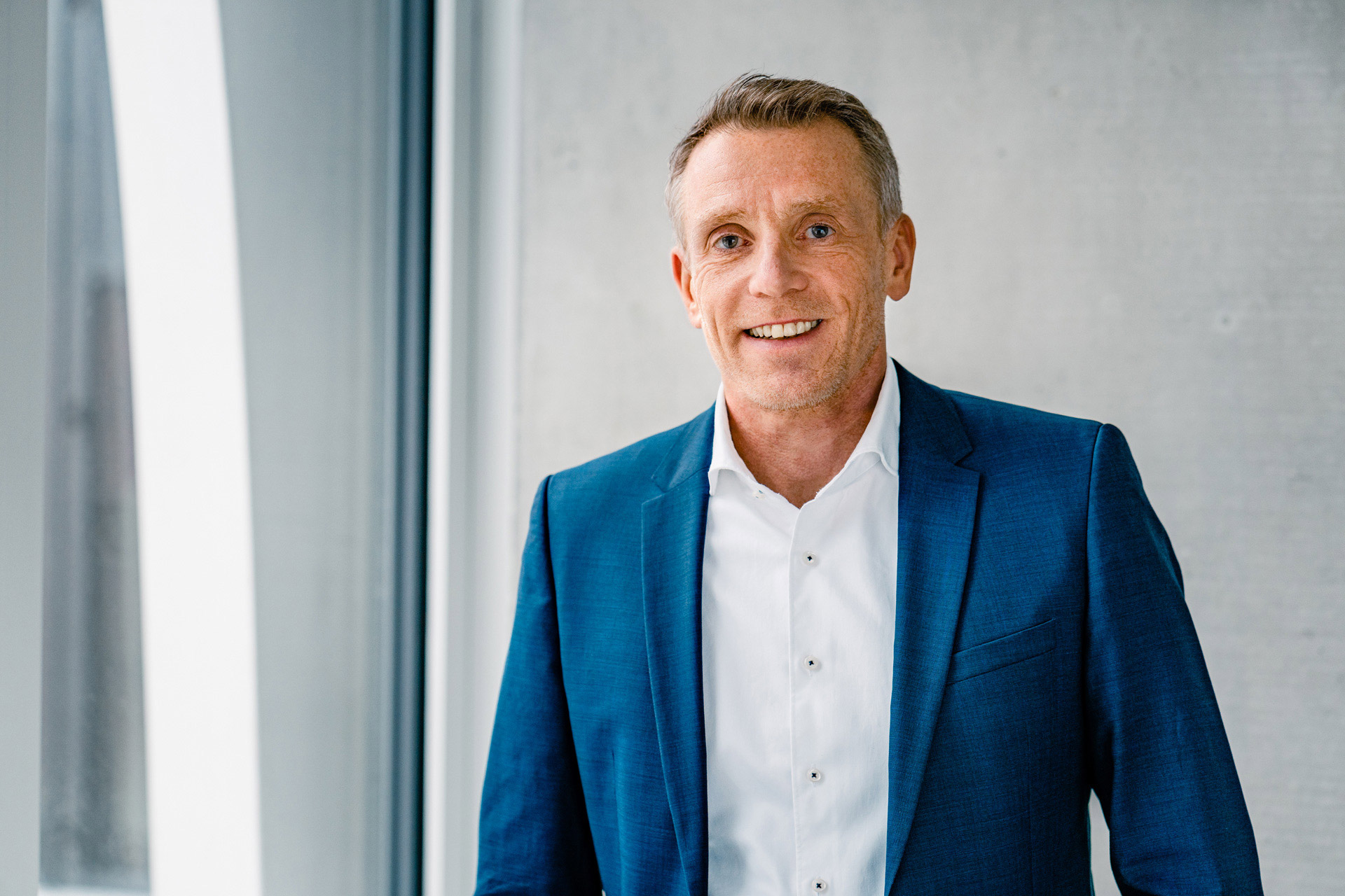 Christoph Beumelburg wearing a white shirt and a blue jacket, in the background a window and an exposed concrete wall