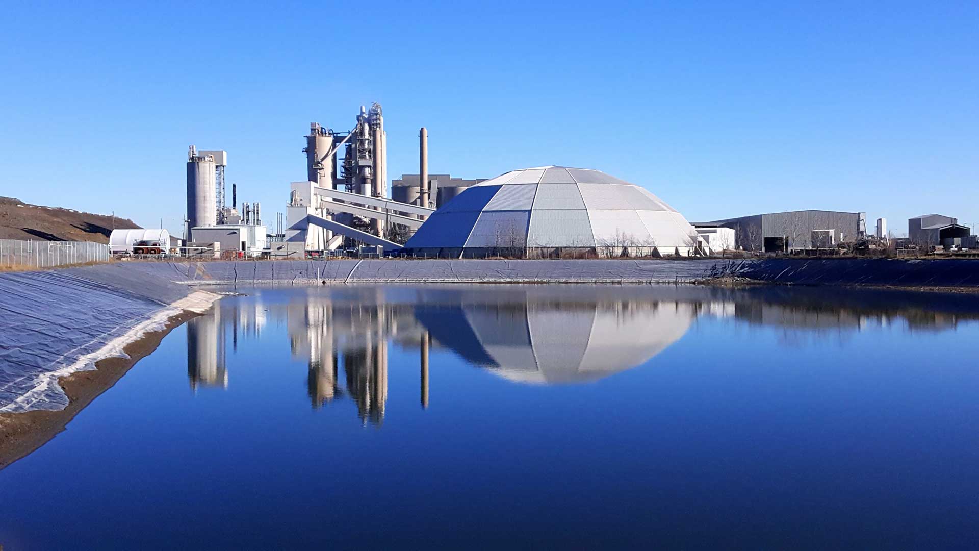 A large cement factory with tall chimneys and a large, dome-shaped building reflected in a body of water. A smaller building is visible to the left.