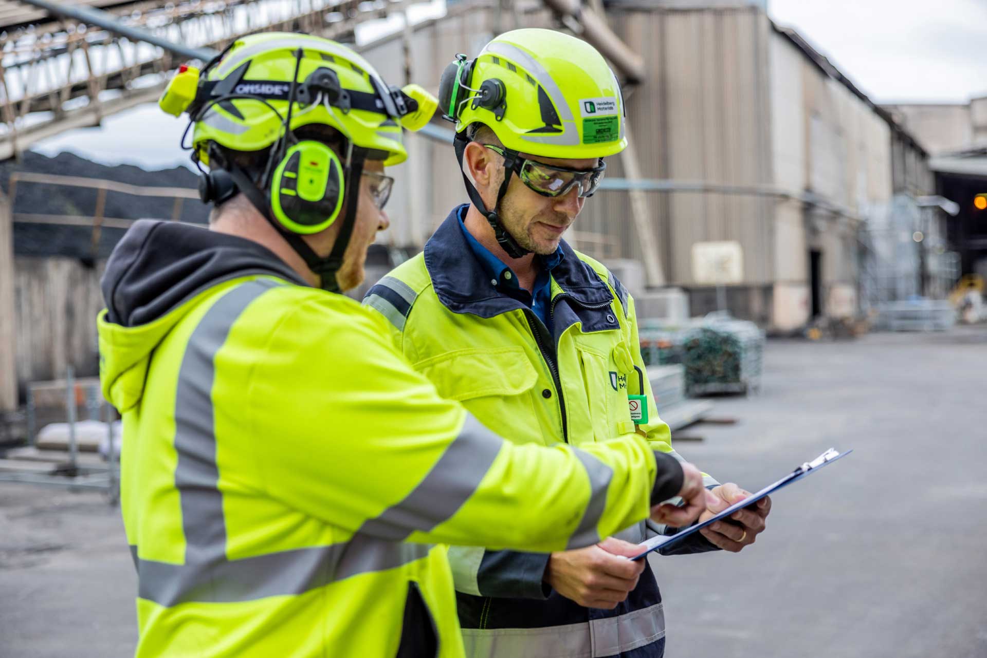 Two employees in protective clothing stand in a factory and look at documents together