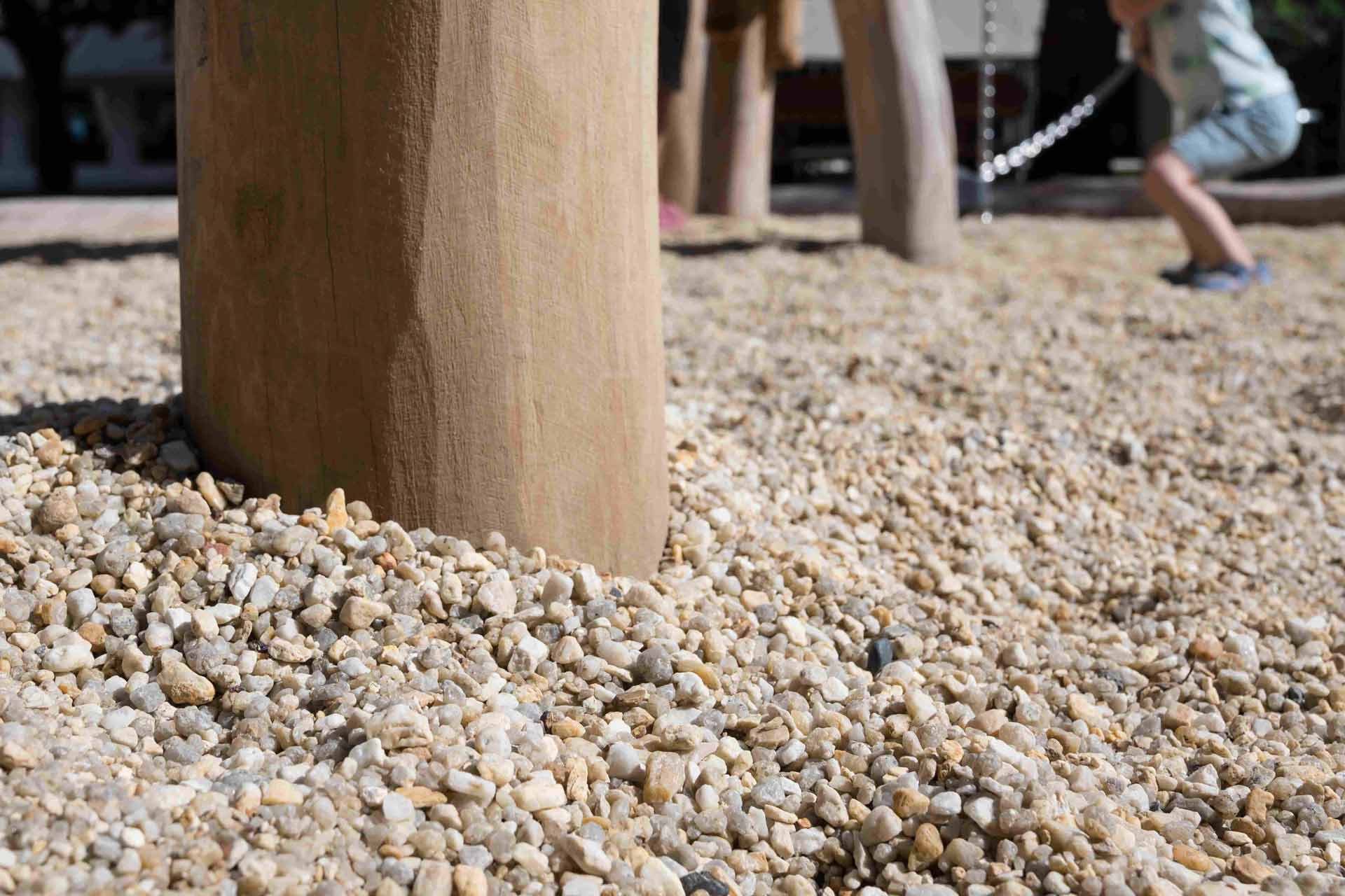 Close-up view of a wooden post planted into the ground, surrounded by light-coloured gravel. In the background, there is a blurred figure of a person engaged in an activity involving a rope or cord.