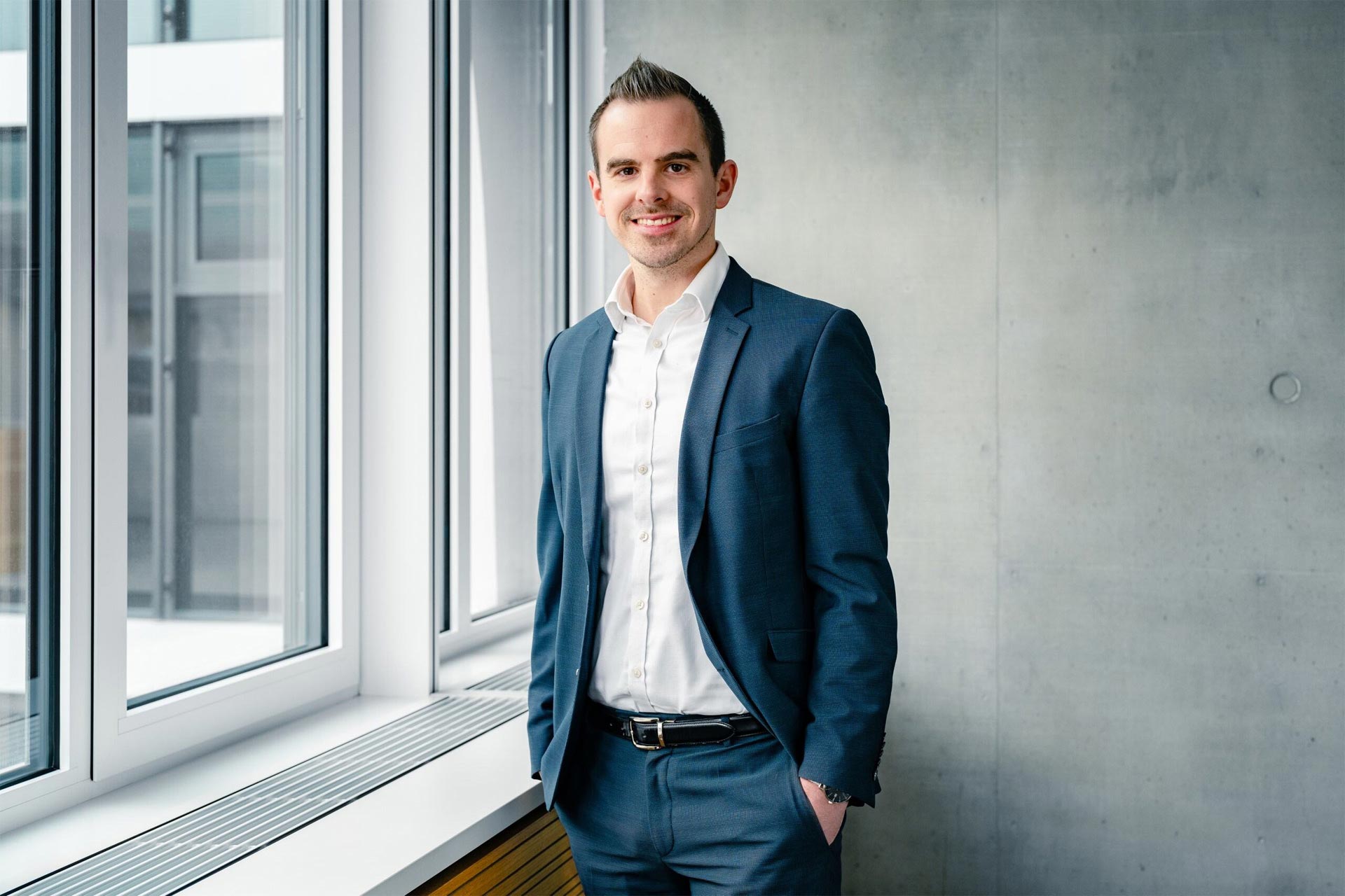 Steven Meyers in a dark blue suit and white shirt in front of an exposed concrete wall, next to a window