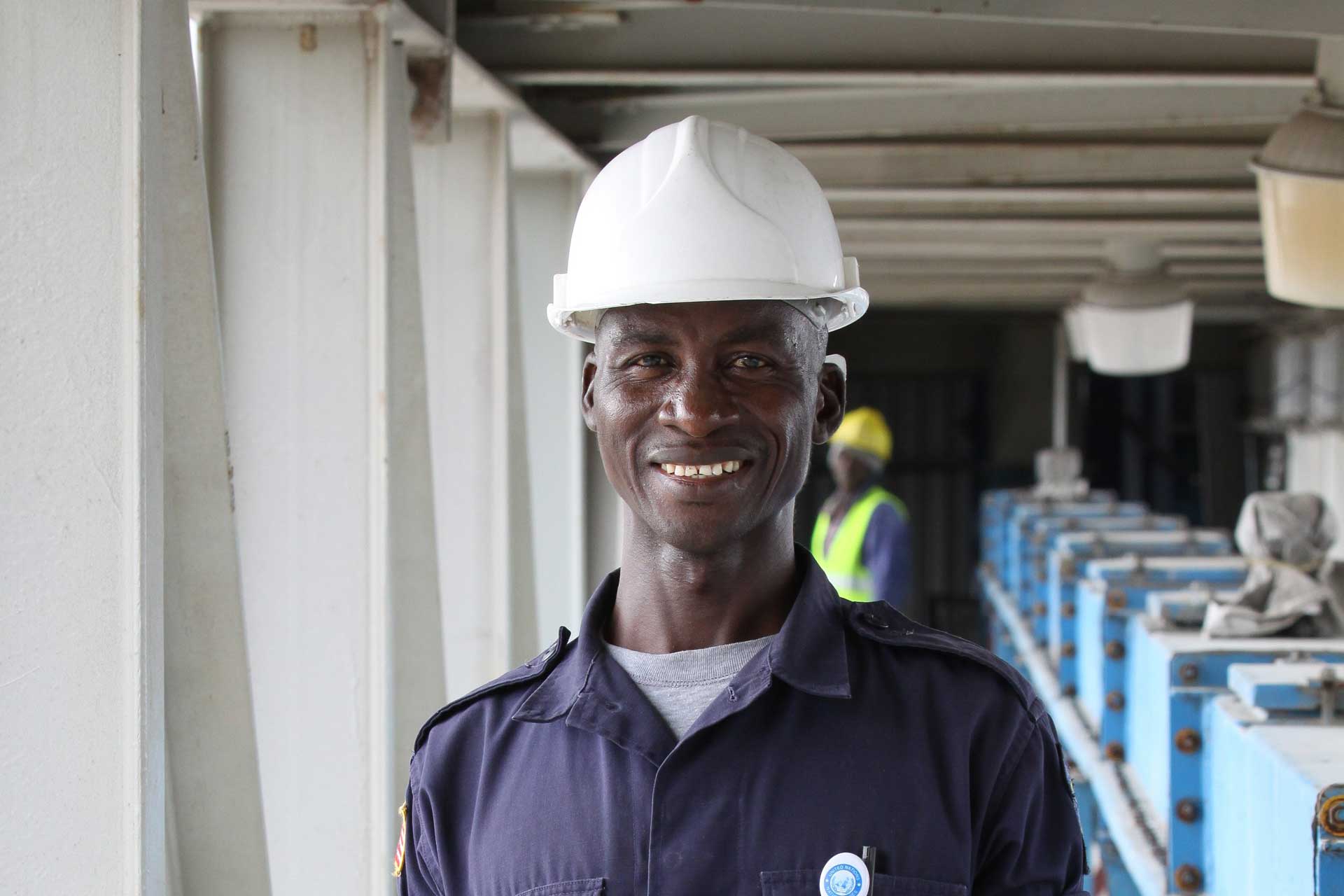 A worker in a blue shirt and white hard hat at a construction site, with another person in the background