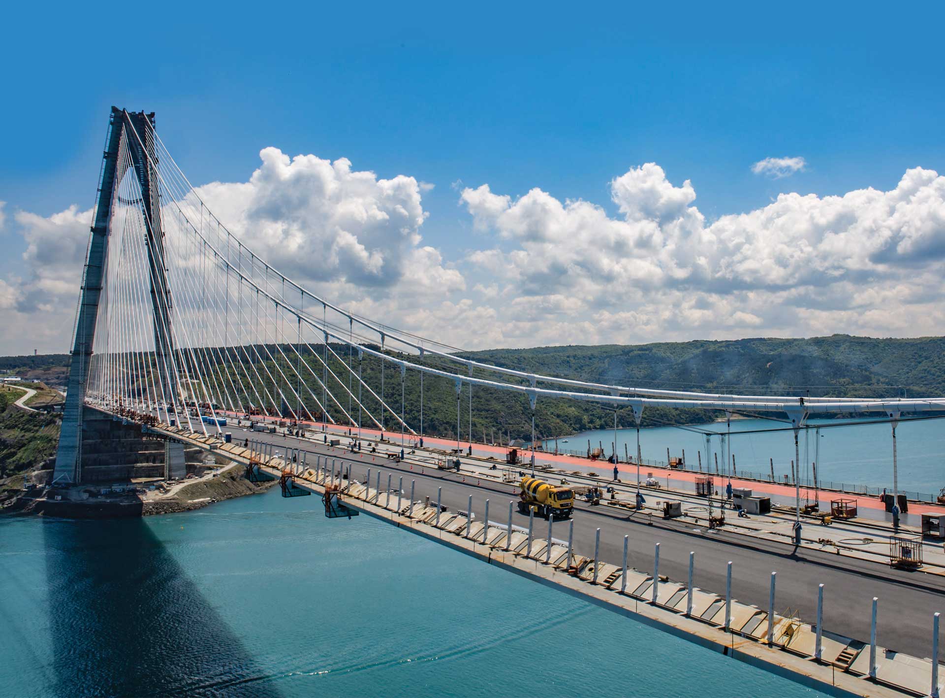 Eine große Hängebrücke im Bau, die ein Gewässer überspannt. Die Brückenfahrbahn ist teilweise fertiggestellt, mit sichtbaren Arbeitern und Baufahrzeugen.