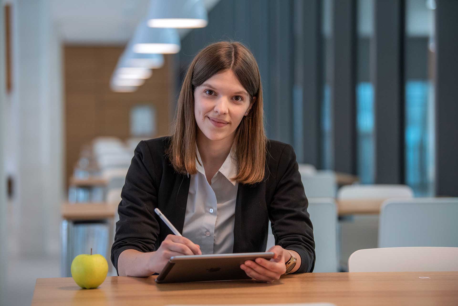 Frau in heller Bluse und dunklem Blazer sitzt an einem Tisch mit einem Tablet, auf dem Tisch liegt ein Apfel