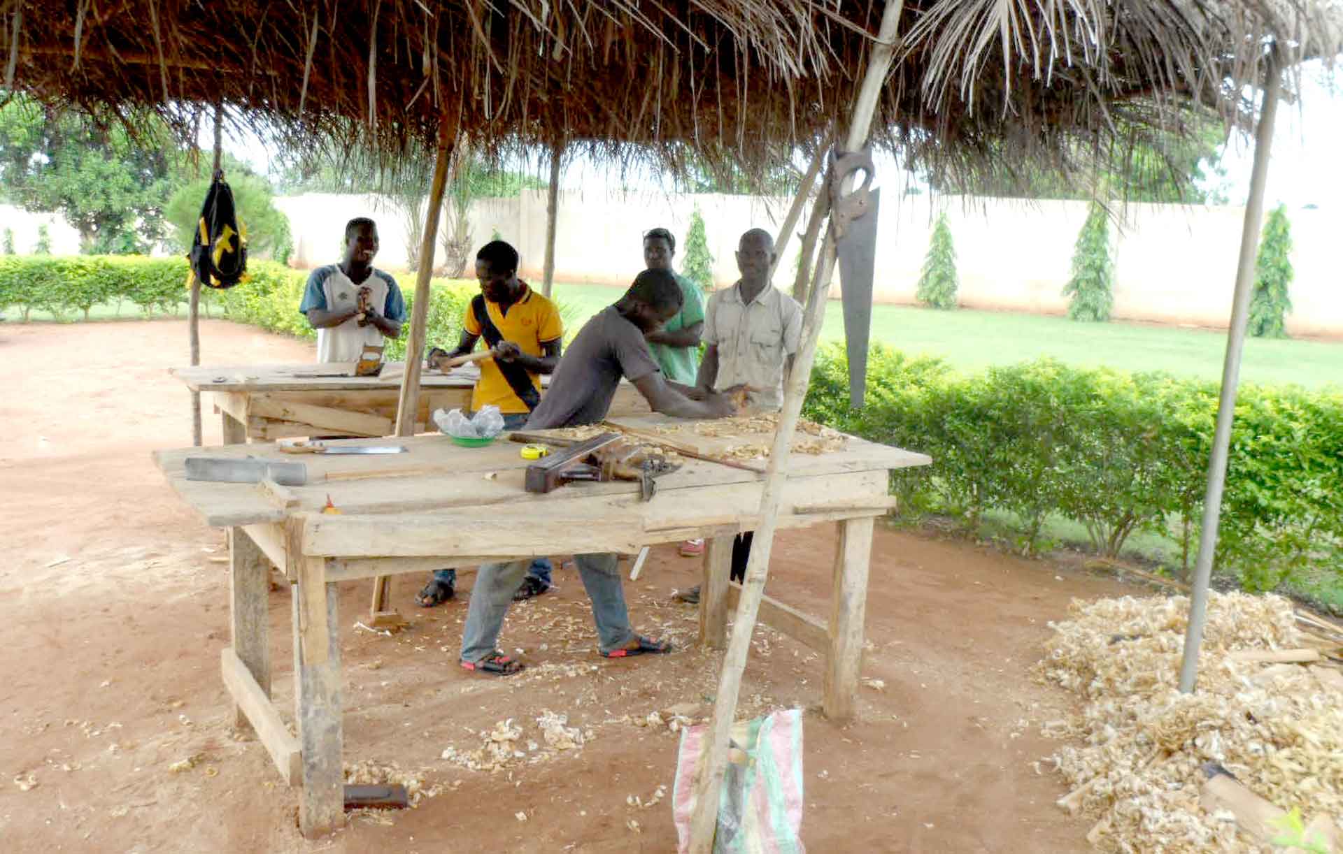 Five men are carpentering two large wooden boards outside, with wood shavings lying on the ground next to them.