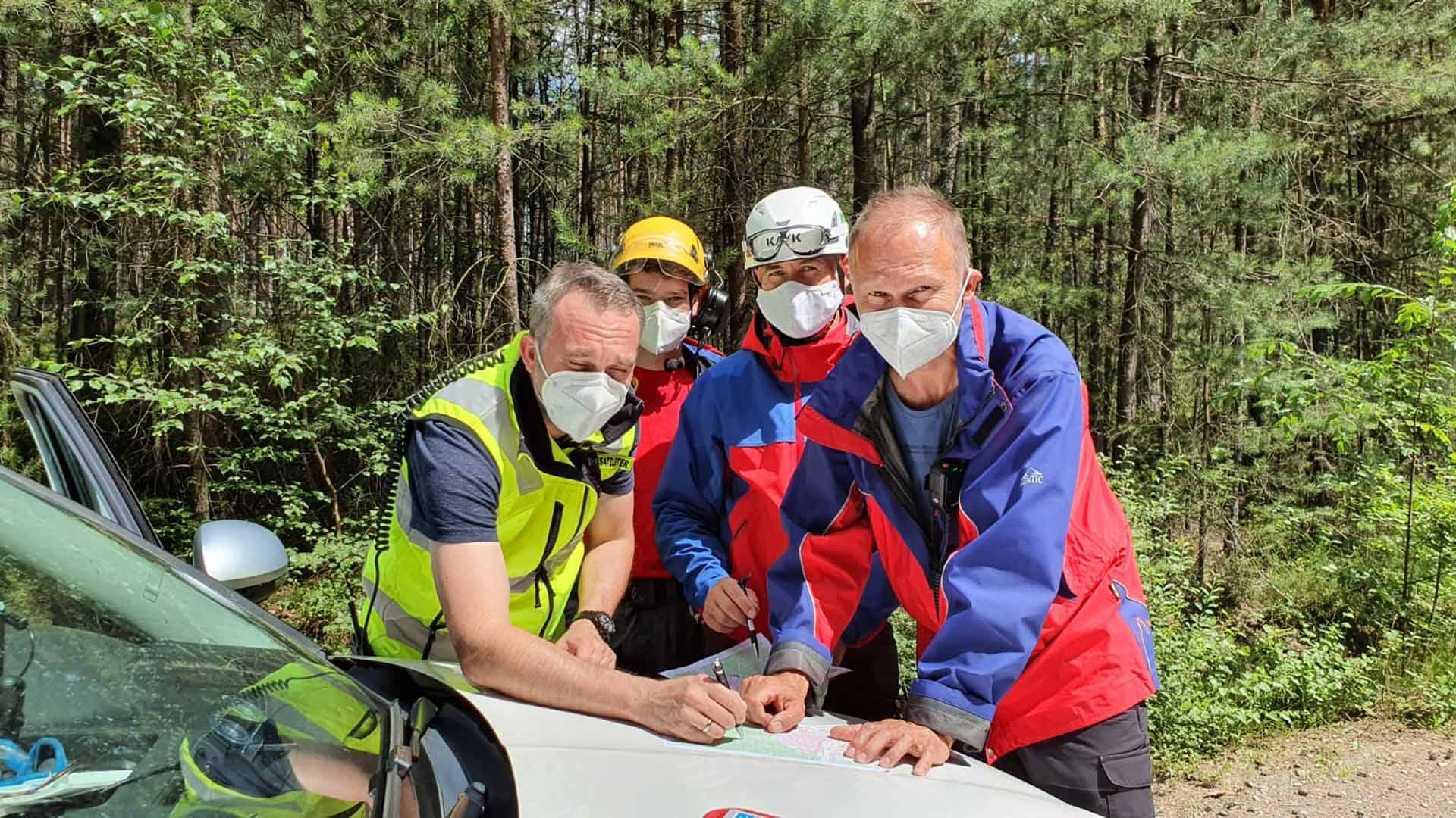 Vier Männer mit FFP2-Masken lehnen an der Motorhaube eines Autos und betrachten eine Karte, im Hintergrund Wald.